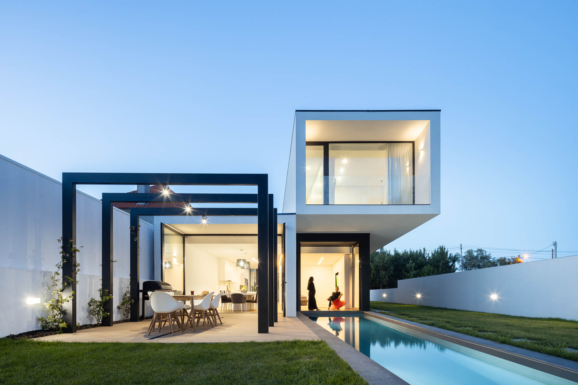 Exterior da casa ao entardecer com piscina iluminada, pérgola e volume branco suspenso. — Casa Diagonal arquitetura Frari em Aveiro