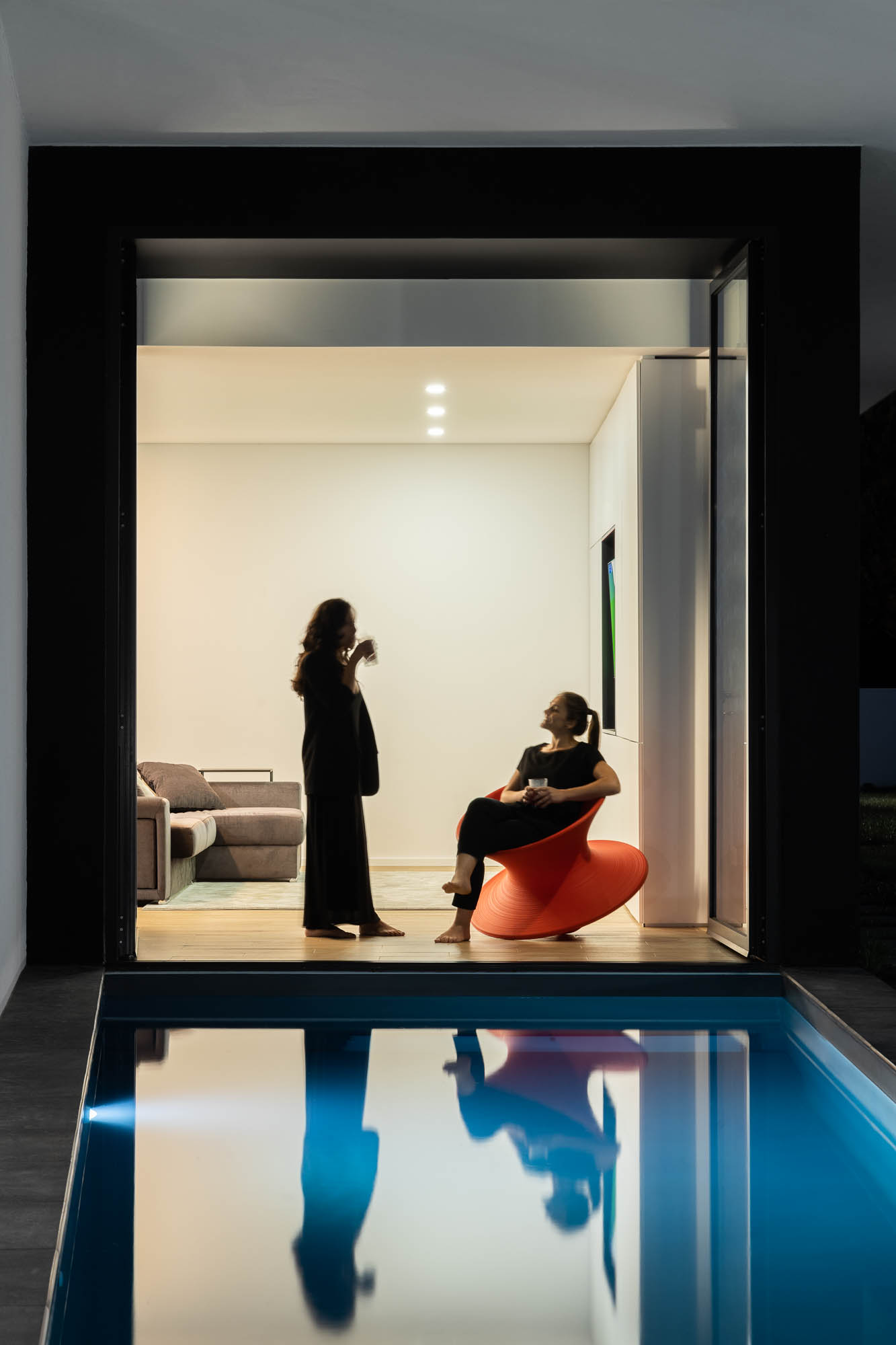 Interior com vista para a piscina, duas mulheres a conversar e reflexos na água. — Casa Diagonal arquitetura Frari em Aveiro