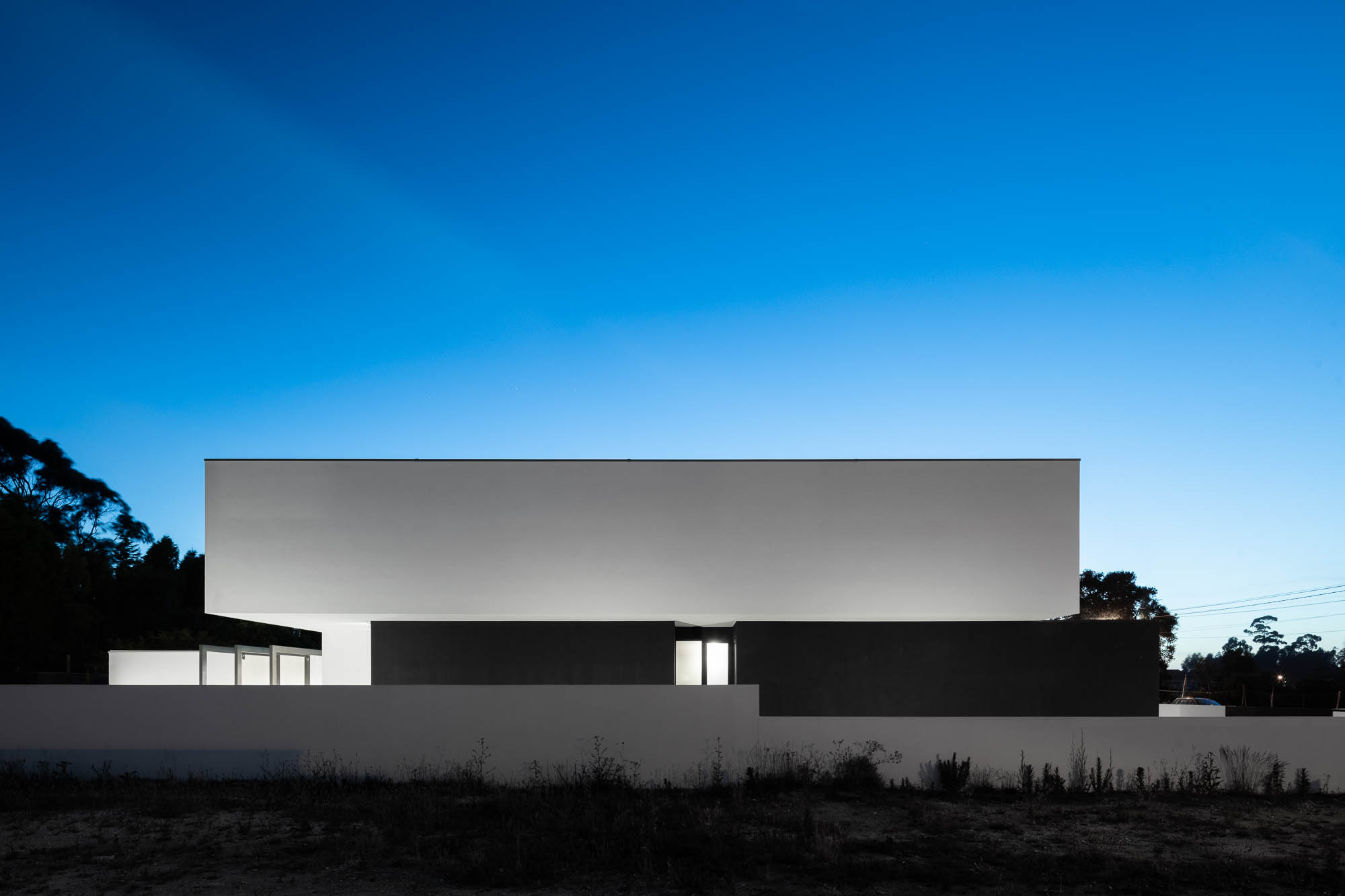 Fachada da casa ao anoitecer com iluminação indireta, muro branco e vegetação ao fundo. — Casa Diagonal arquitetura Frari em Aveiro