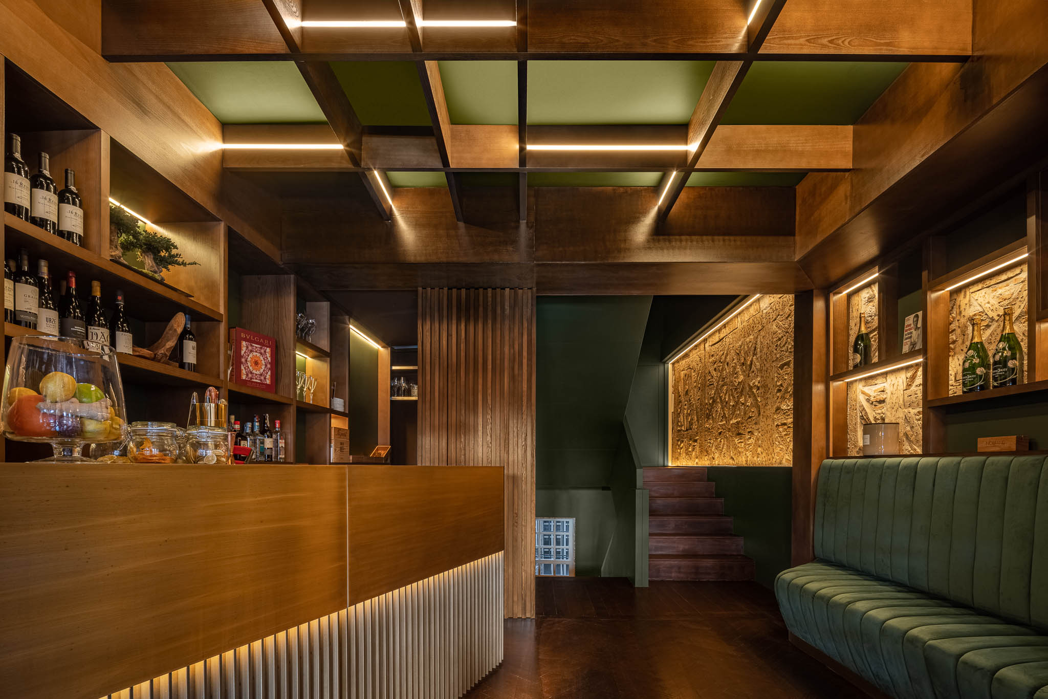 Balcão de madeira clara com iluminação LED, prateleiras com garrafas e um sofá verde escuro ao fundo. — Restaurante 34 arquitetura REM'A Arquitectos em Guimarães