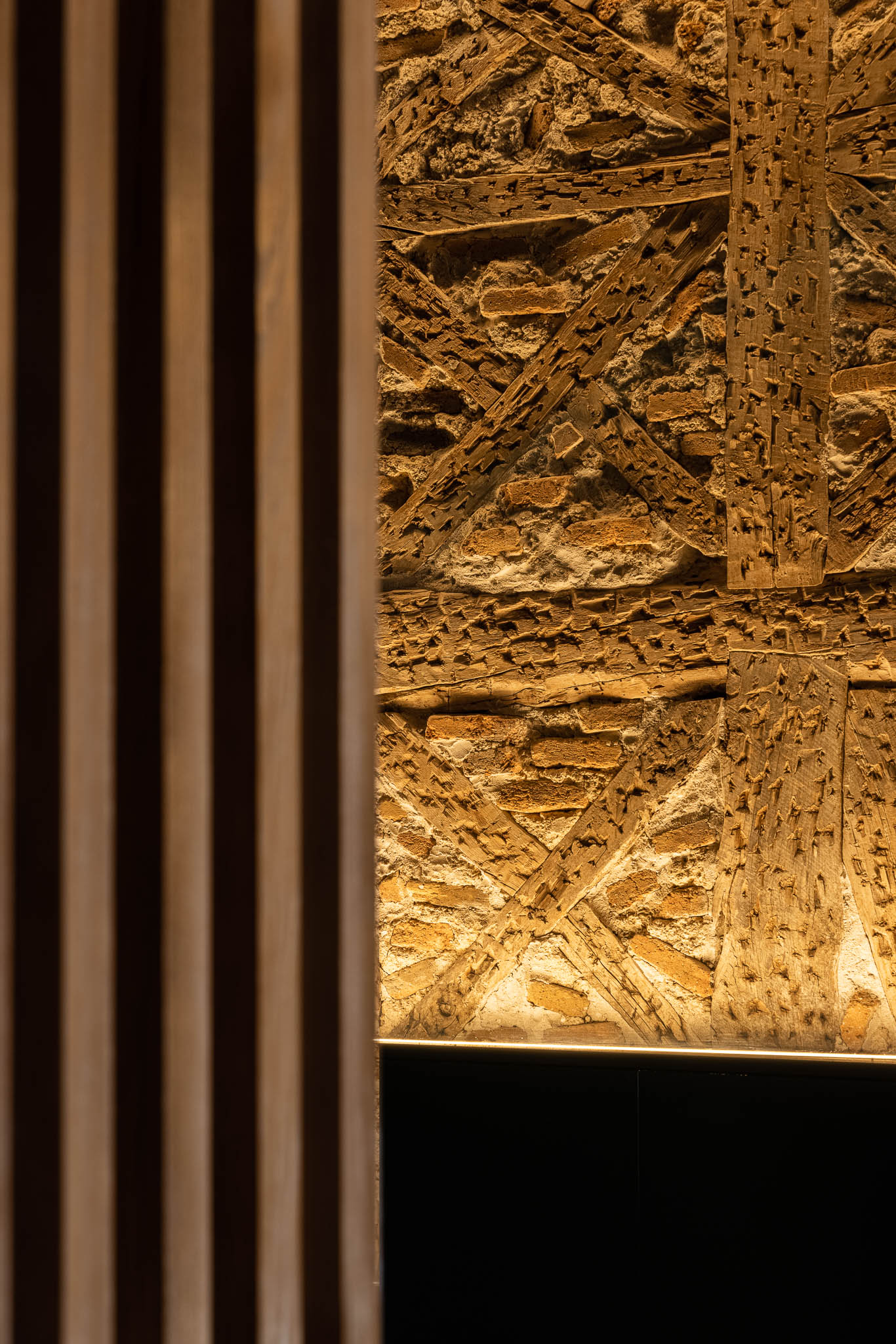 Pormenor de uma parede com tijolos e vigas de madeira expostas, iluminada, ao lado de ripas verticais de madeira. — Restaurante 34 arquitetura REM'A Arquitectos em Guimarães