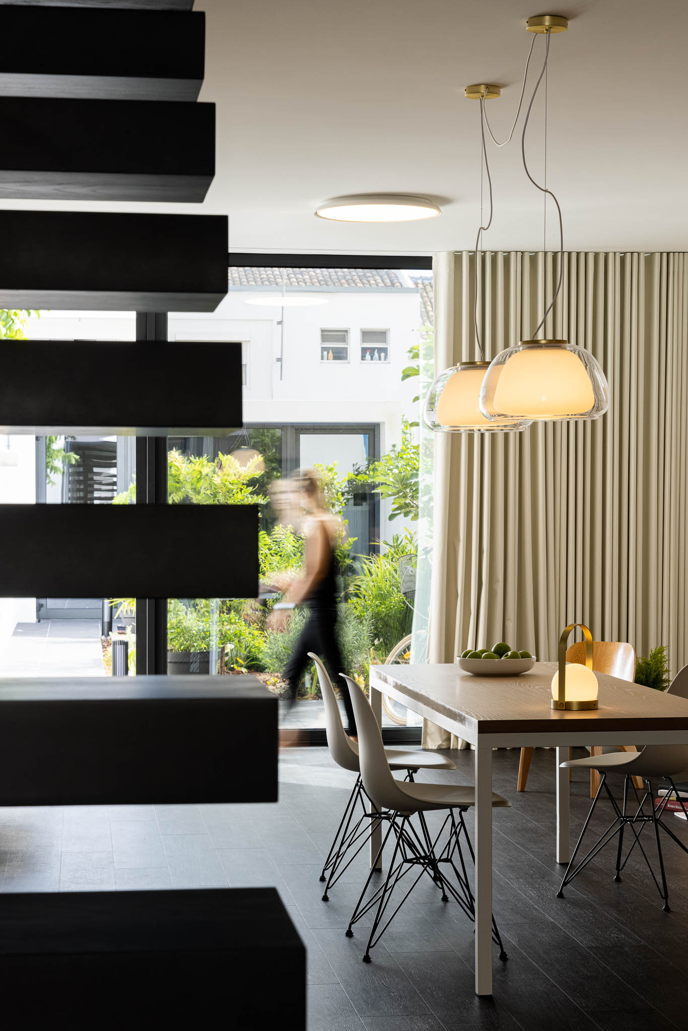 Sala de jantar com escada preta em primeiro plano, mesa de madeira clara e cadeiras brancas. Iluminação suspensa moderna. — Casa moinho de vento arquitetura BOX arquitectos em Ponta Delgada