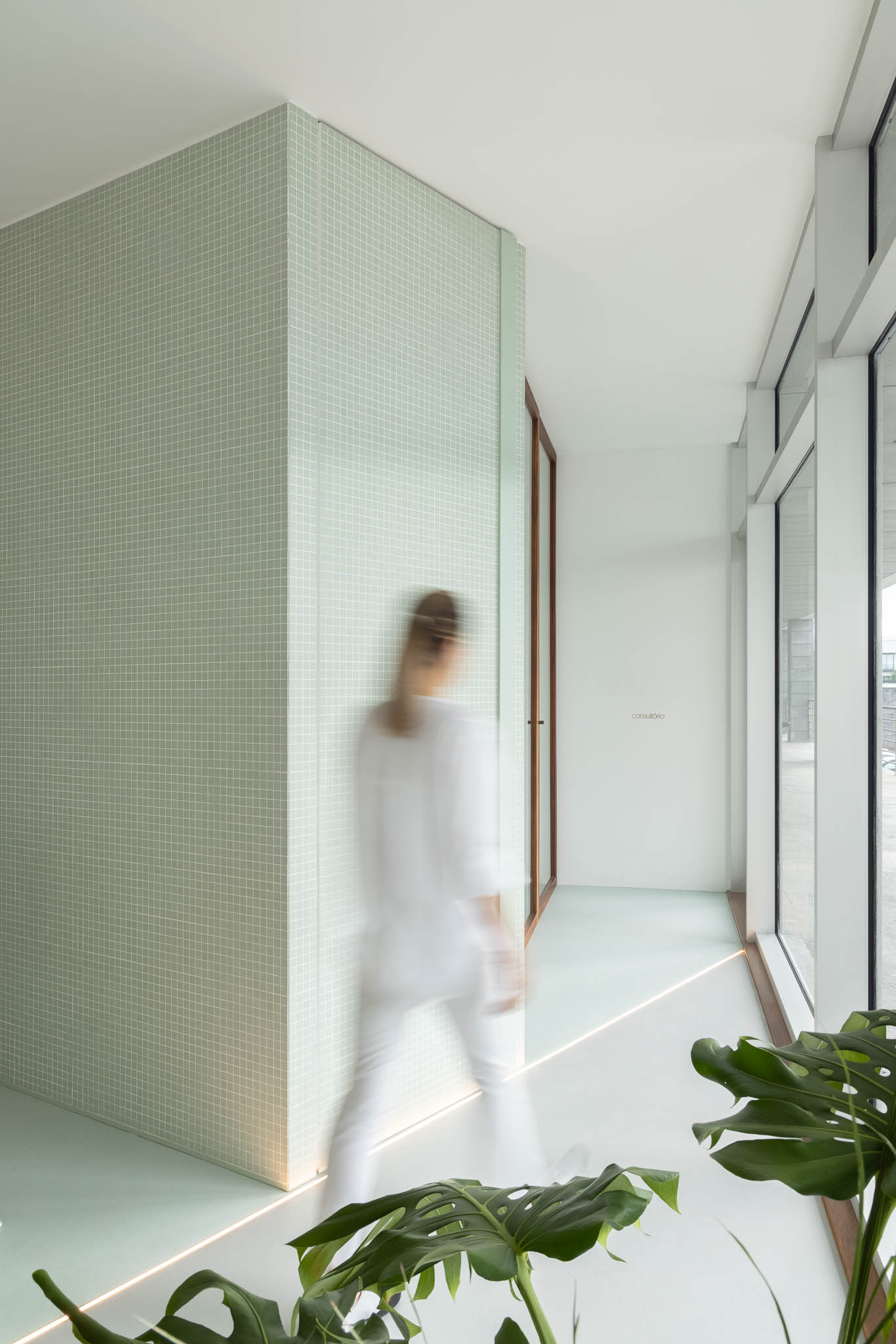 Corredor com azulejos verde-água, iluminação embutida no chão e grandes janelas, criando uma atmosfera calma e luminosa. — Think Health Naturopatia arquitetura BOX arquitectos em Ponta Delgada