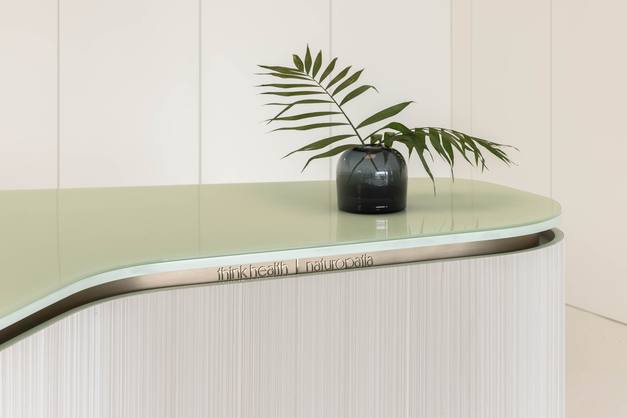 Detalhe do balcão de receção com tampo de vidro, vaso com planta e logótipo "think health | naturopatia", reforçando a identidade da marca. — Think Health Naturopatia arquitetura BOX arquitectos em Ponta Delgada