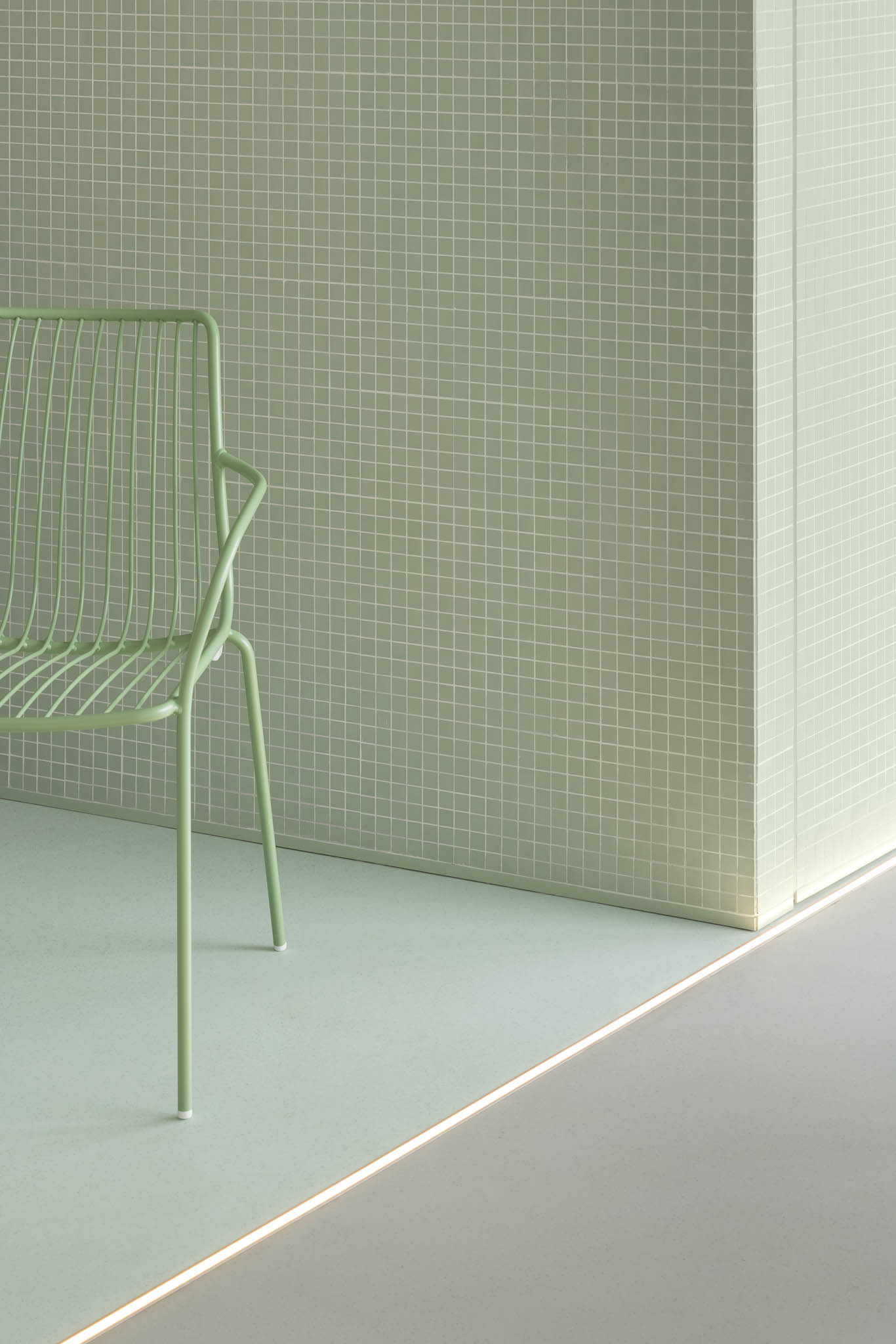 Detalhe de uma cadeira metálica verde-água junto a uma parede revestida com azulejos do mesmo tom, com iluminação embutida no chão. — Think Health Naturopatia arquitetura BOX arquitectos em Ponta Delgada