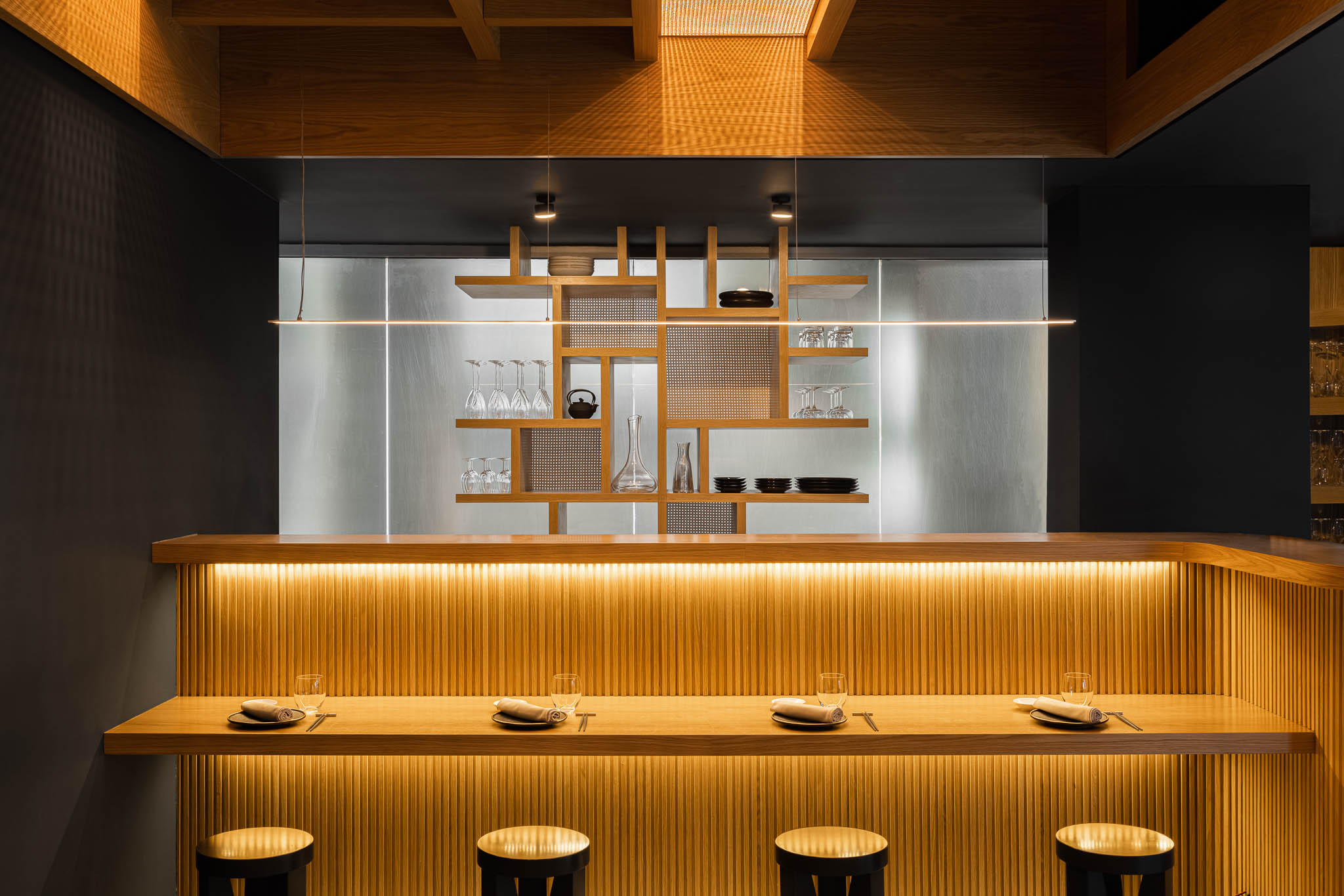 Balcão de bar em madeira clara com iluminação embutida, cadeiras pretas e prateleiras com louça, num ambiente minimalista e elegante. — Restaurante Fuji arquitetura Sequeira Dias Arquitectos em Ponta Delgada
