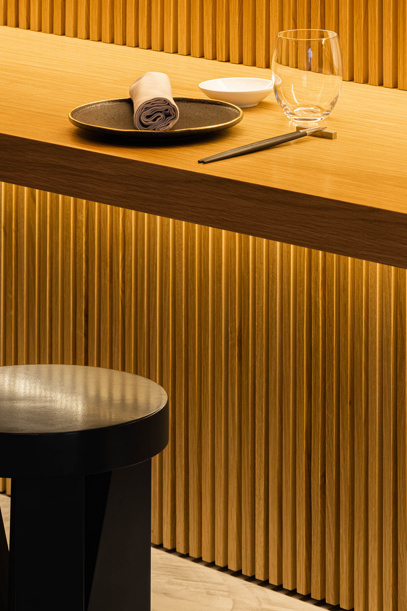 Close-up do balcão em madeira com iluminação, mostrando a mise en place com prato, guardanapo, pauzinhos e copo. — Restaurante Fuji arquitetura Sequeira Dias Arquitectos em Ponta Delgada