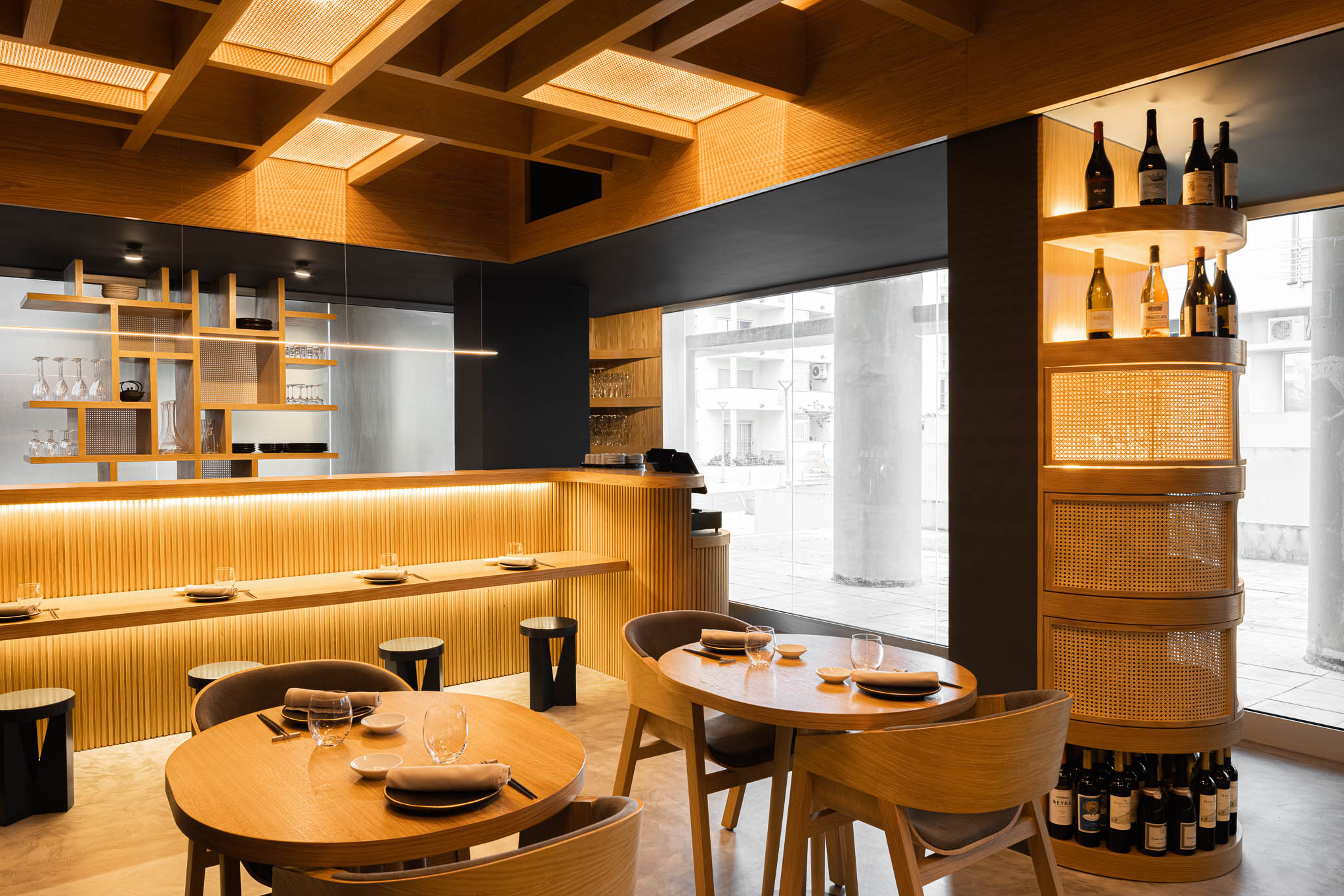 Interior do restaurante com mesas, cadeiras em madeira clara, balcão iluminado e garrafeira, com luz natural ao fundo. — Restaurante Fuji arquitetura Sequeira Dias Arquitectos em Ponta Delgada
