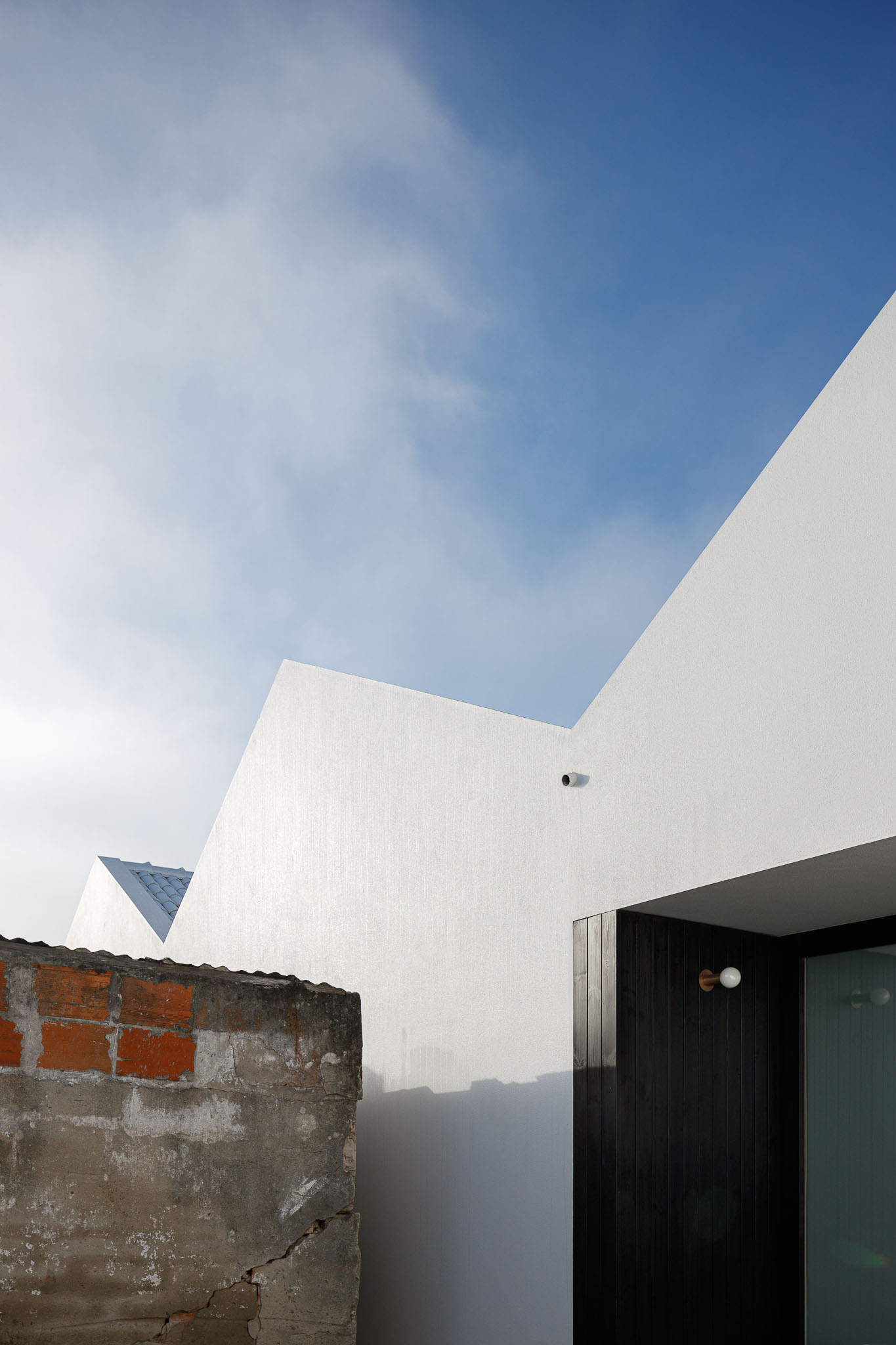 Fachada branca minimalista com telhados triangulares, detalhe de madeira escura e parede rústica em tijolo e betão. — Habitação em Santa Joana arquitetura Nu.ma em Aveiro