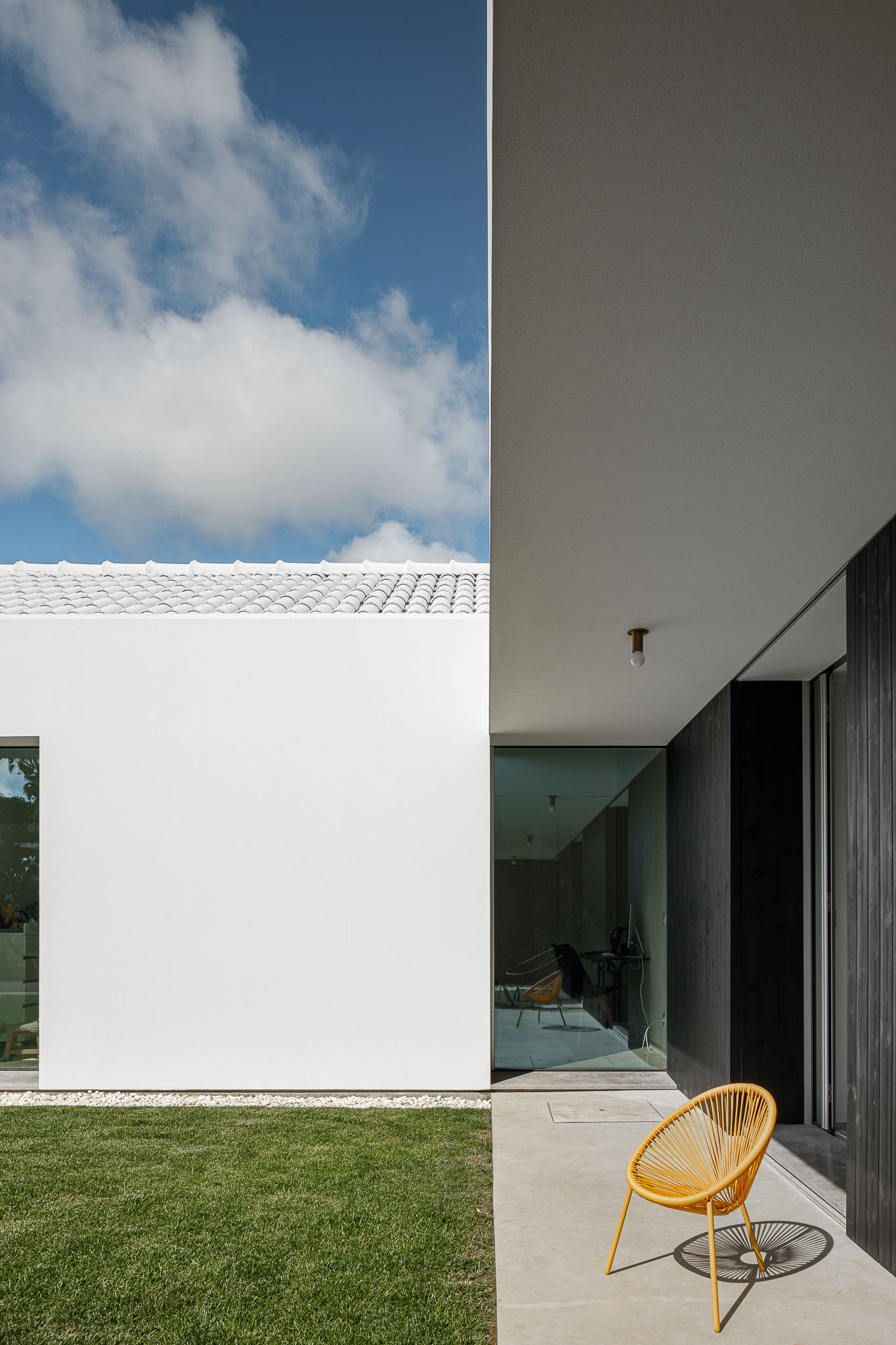 Detalhe exterior com fachada branca, revestimento em madeira escura e cadeira amarela no pátio. — Habitação em Santa Joana arquitetura Nu.ma em Aveiro