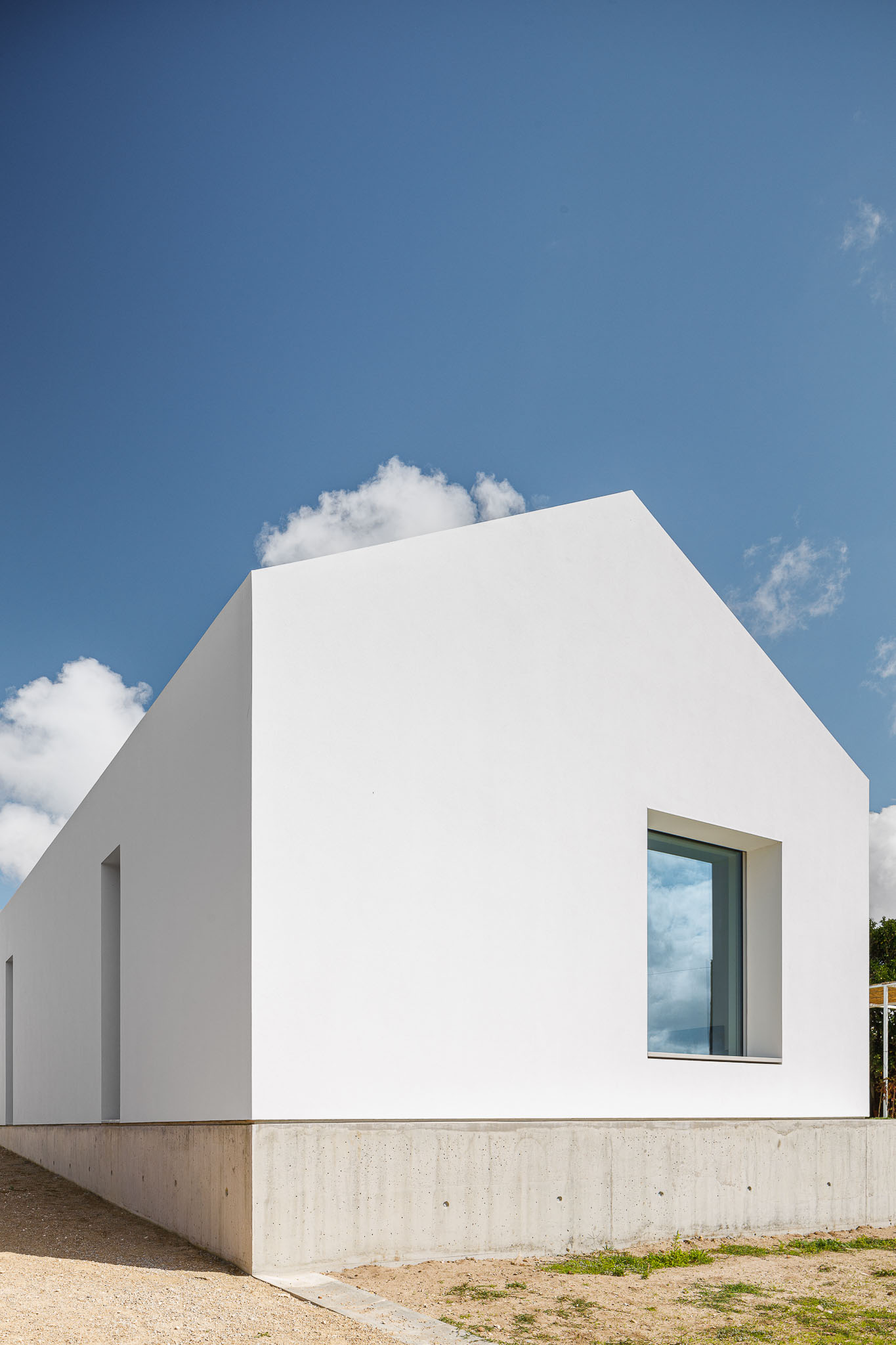 Fachada lateral da casa com volume branco puro sobre base de betão aparente e céu azul. — Habitação em Santa Joana arquitetura Nu.ma em Aveiro