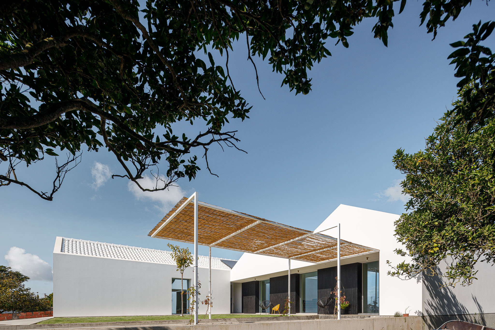 Vista exterior da casa com alpendre de bambu, volumes brancos e vegetação envolvente. — Habitação em Santa Joana arquitetura Nu.ma em Aveiro