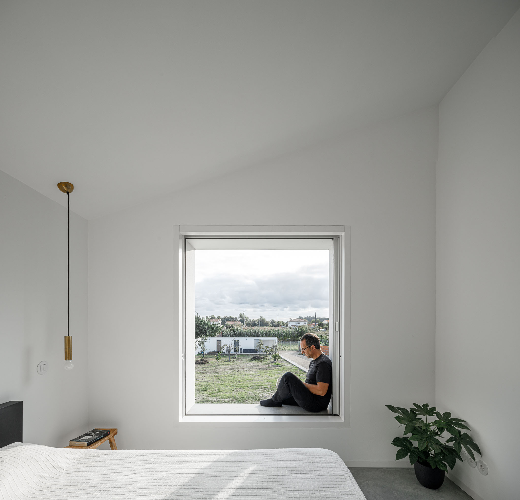 Quarto com janela ampla, homem sentado, cama branca e pavimento em betão polido. — Habitação em Santa Joana arquitetura Nu.ma em Aveiro