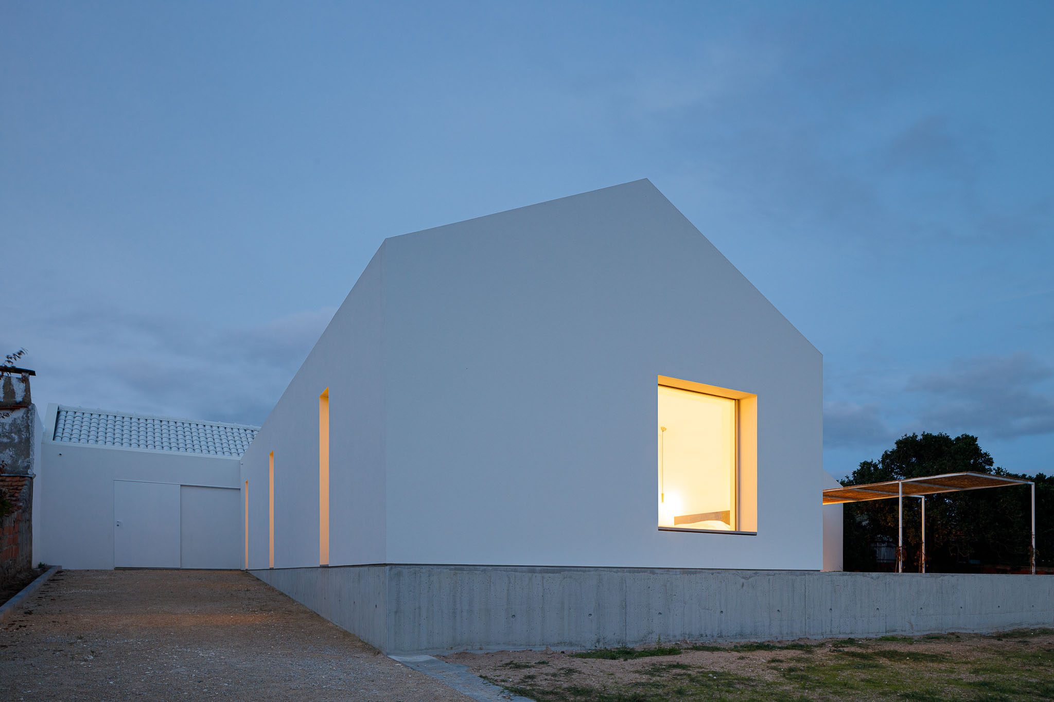 Vista noturna da casa com volumes brancos iluminados, base em betão e telhado branco. — Habitação em Santa Joana arquitetura Nu.ma em Aveiro