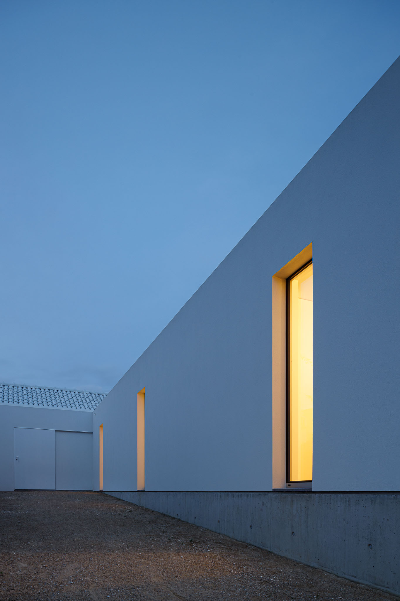 Detalhe noturno da fachada branca com janelas iluminadas e telhado branco ao fundo. — Habitação em Santa Joana arquitetura Nu.ma em Aveiro