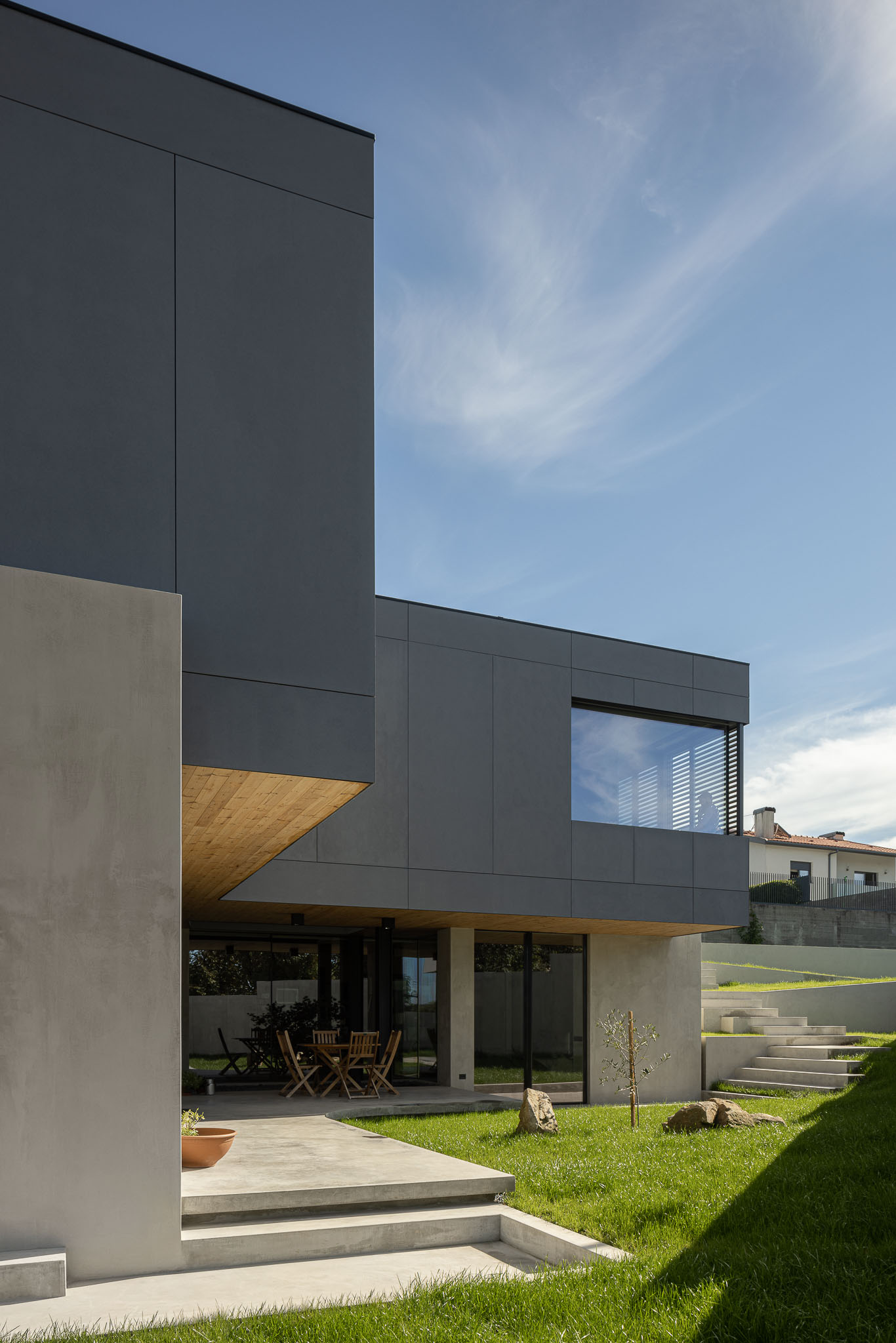 Fachada da casa com revestimento cinzento e detalhes em madeira, jardim relvado e escadas de acesso em betão. — Casa 15 arquitetura AM-arqstudio em Braga