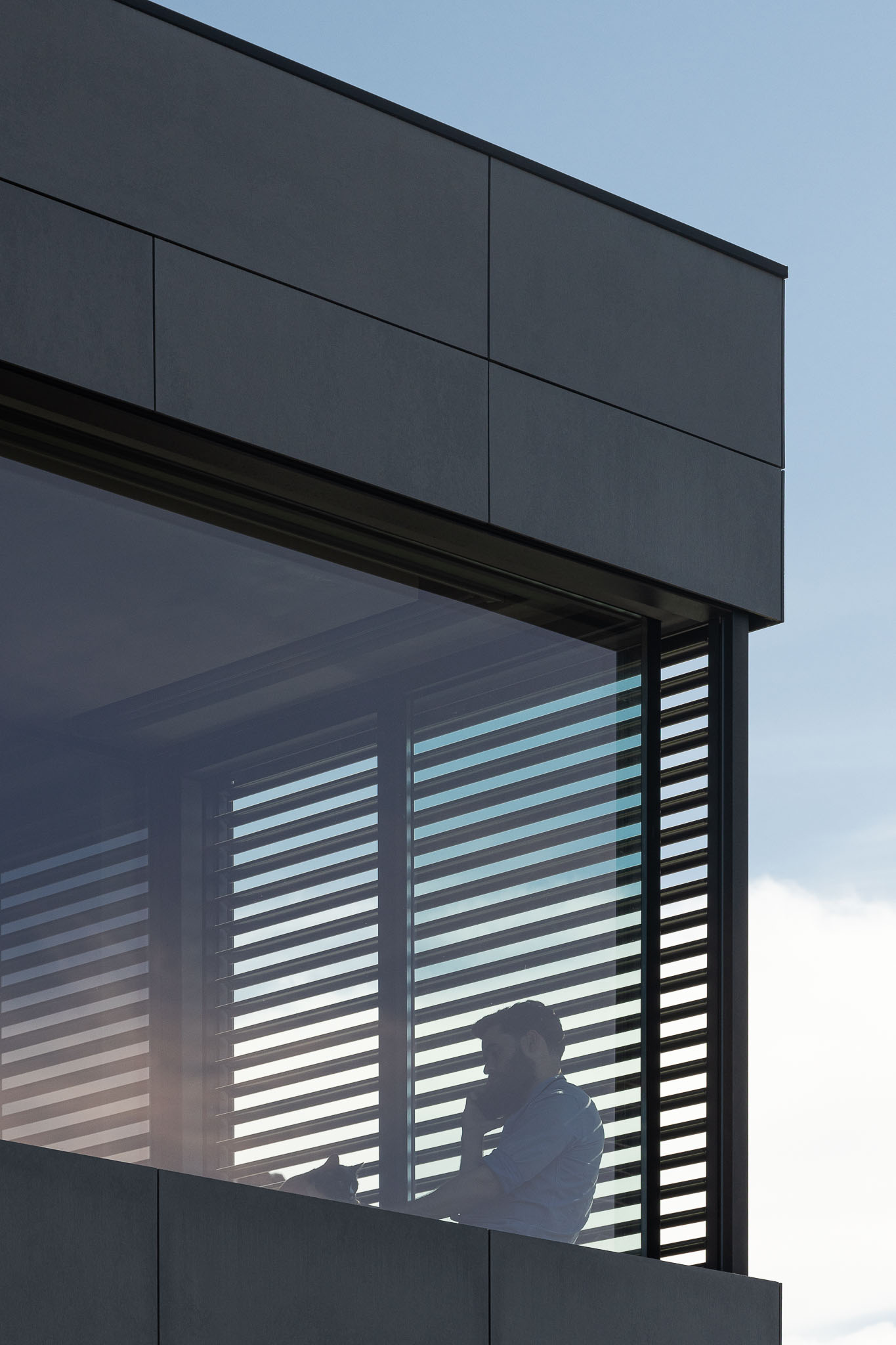 Detalhe da janela com persianas horizontais, fachada cinzenta e céu azul. Homem ao telefone no interior. — Casa 15 arquitetura AM-arqstudio em Braga