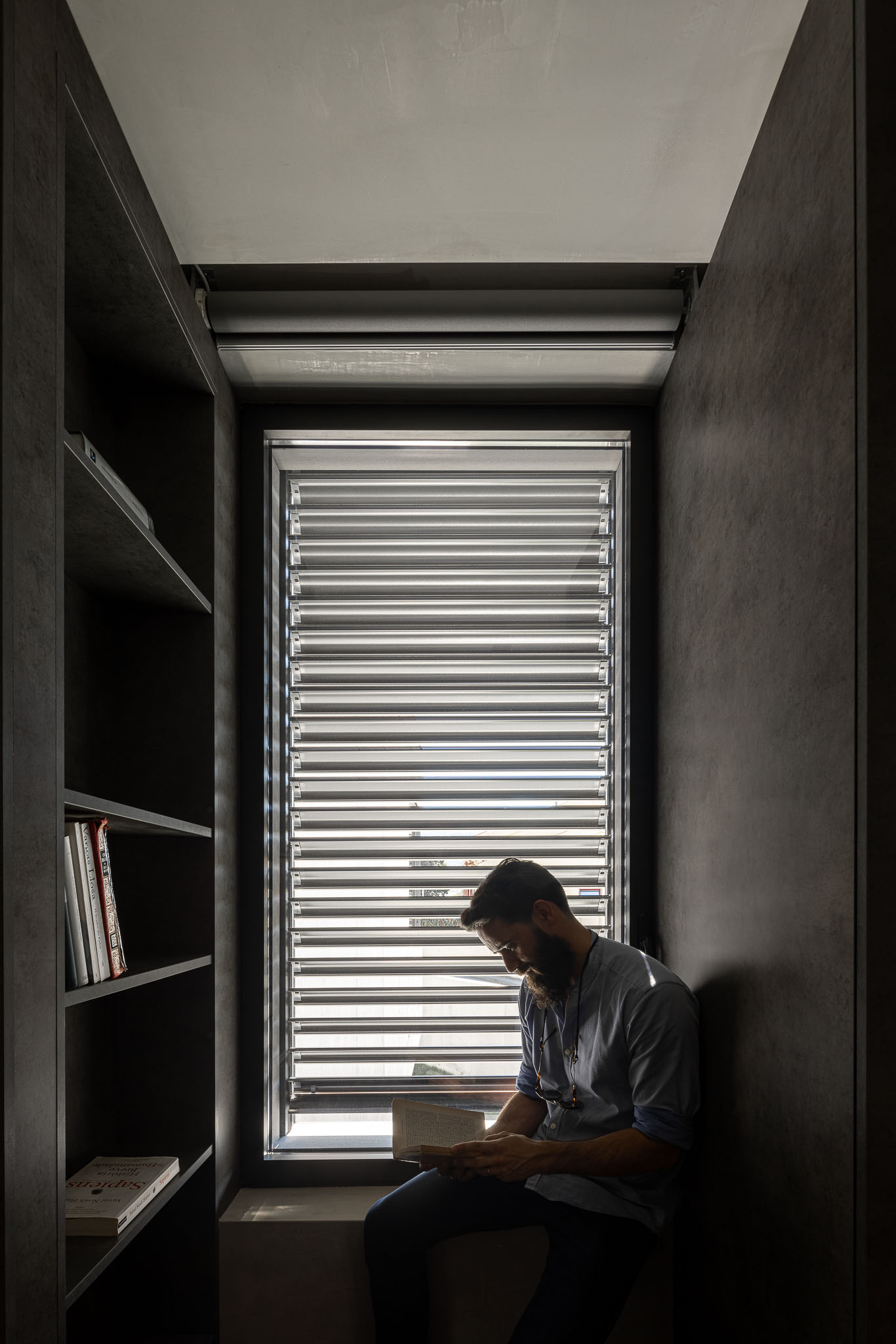 Espaço de leitura com estantes escuras, persianas horizontais e homem a ler junto à janela. — Casa 15 arquitetura AM-arqstudio em Braga
