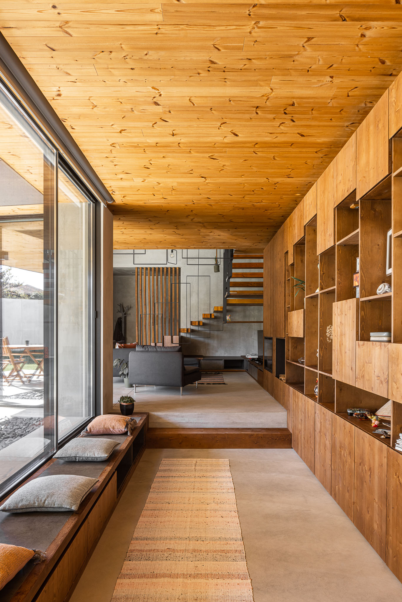 Sala de estar com estante em madeira, sofá cinzento e vista para o exterior através de janelas amplas. — Casa 15 arquitetura AM-arqstudio em Braga