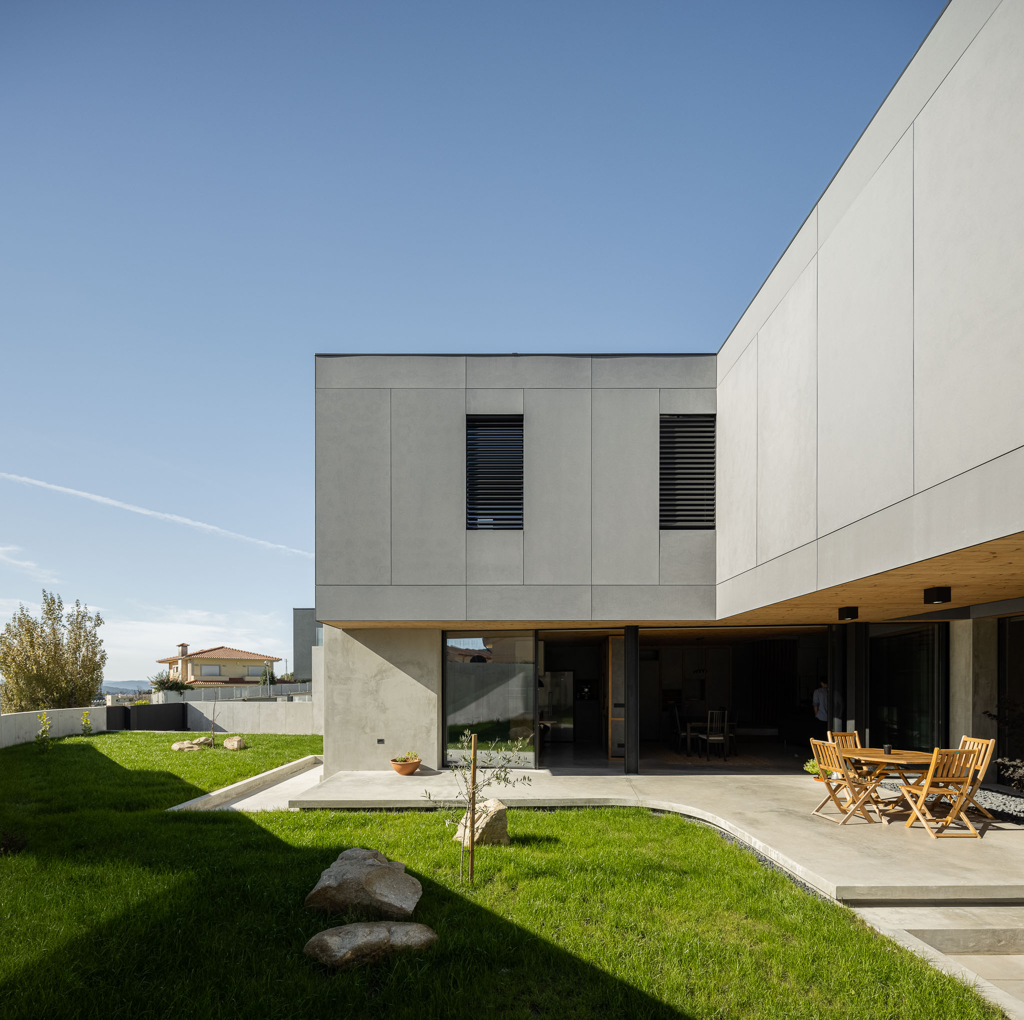 Fachada da casa com revestimento cinzento, jardim relvado e zona de estar exterior com mobiliário em madeira. — Casa 15 arquitetura AM-arqstudio em Braga