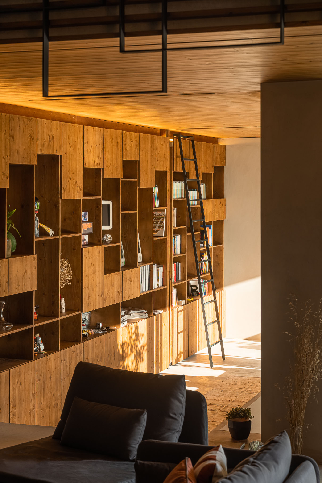 Sala de estar com estante em madeira, sofá cinzento e escada de acesso ao piso superior. — Casa 15 arquitetura AM-arqstudio em Braga