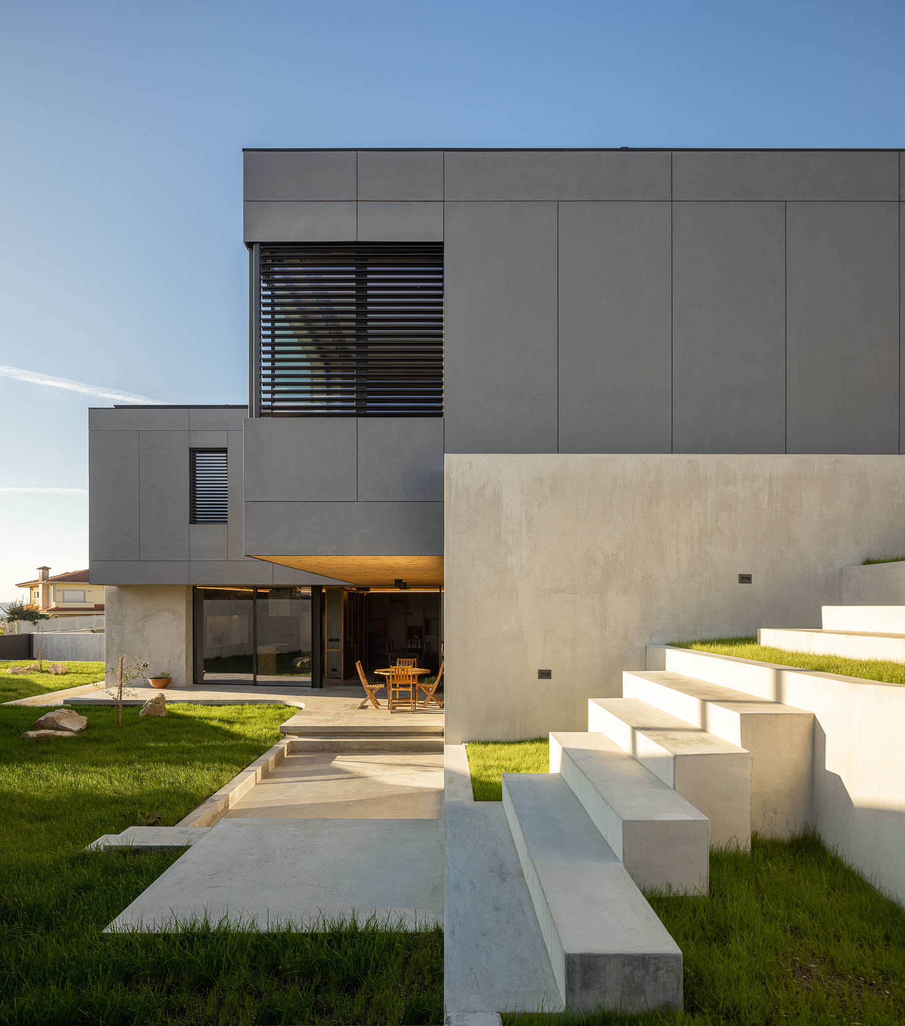 Fachada da casa com revestimento cinzento, jardim relvado e escadas de acesso em betão. — Casa 15 arquitetura AM-arqstudio em Braga