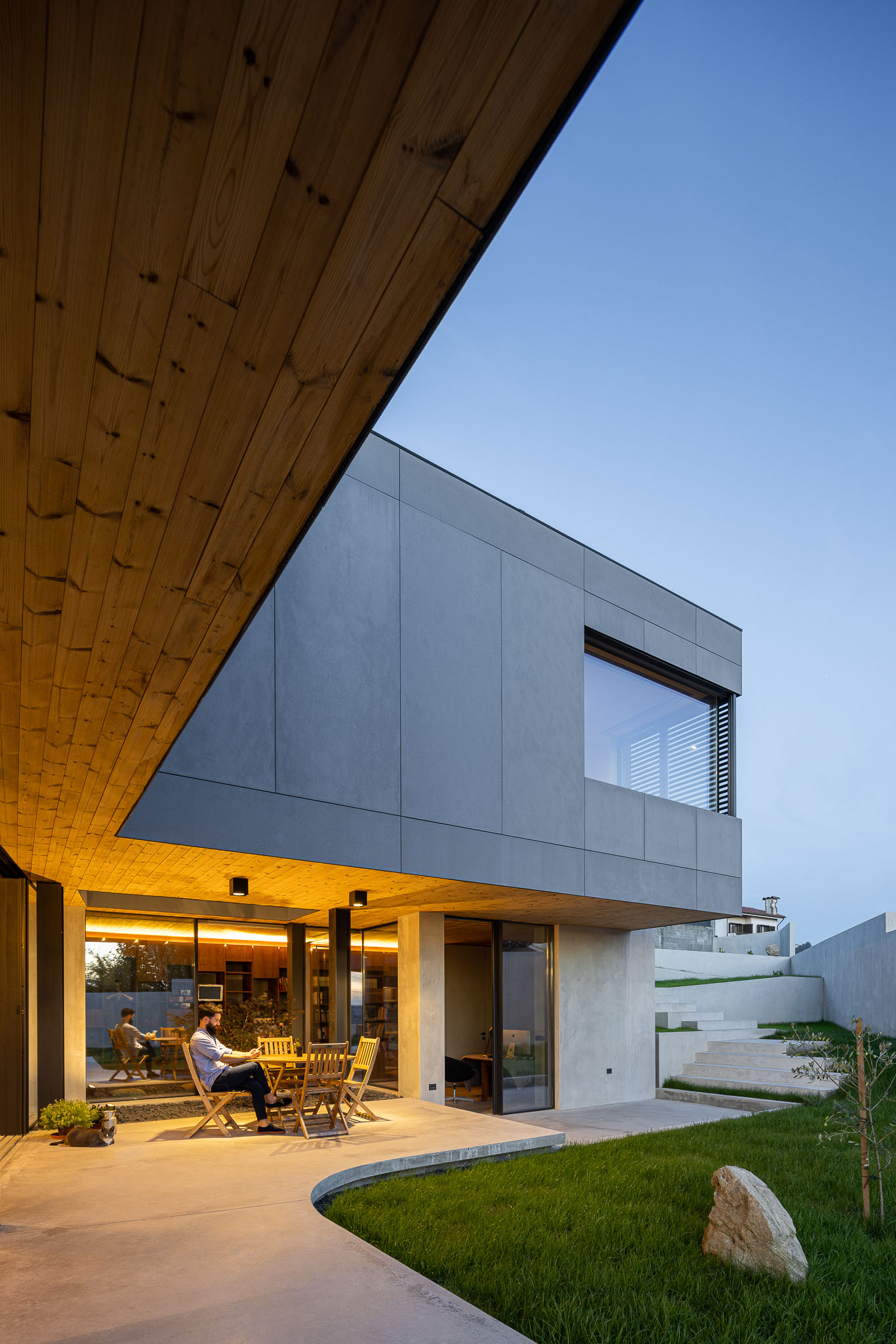 Fachada da casa com revestimento cinzento, detalhes em madeira e zona de estar exterior iluminada. — Casa 15 arquitetura AM-arqstudio em Braga