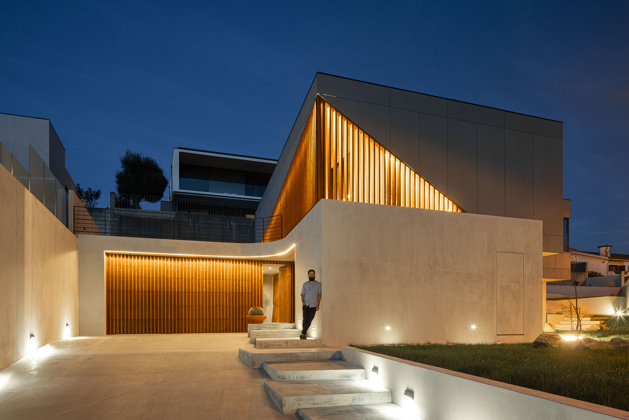 Fachada da casa iluminada à noite, com revestimento cinzento e detalhes em madeira. — Casa 15 arquitetura AM-arqstudio em Braga