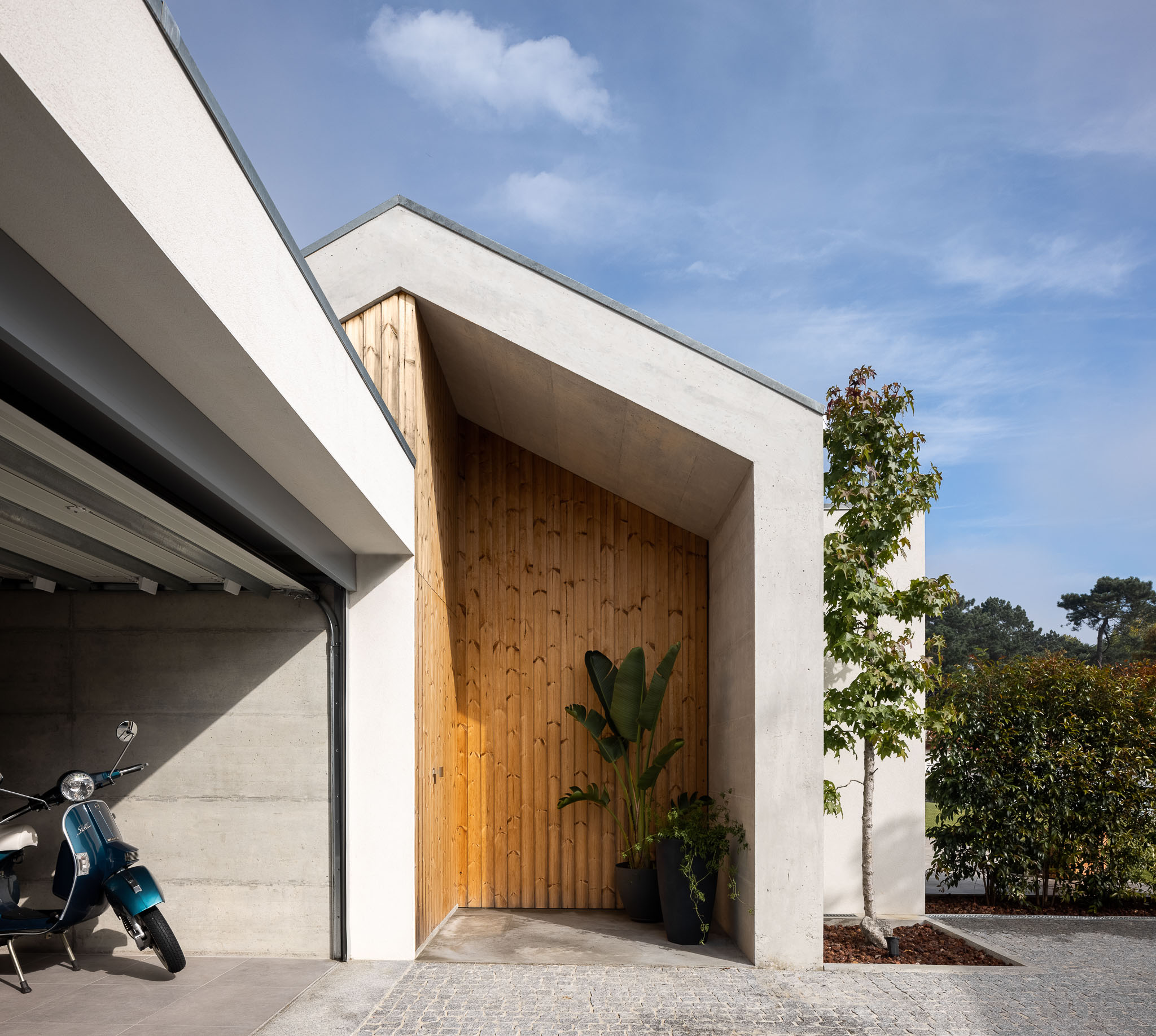 Entrada da casa com porta de madeira vertical, garagem com Vespa azul, paredes em betão aparente e reboco branco. — Casa H1 arquitetura Bruno H Gomes Arquitectura em São João da Madeira