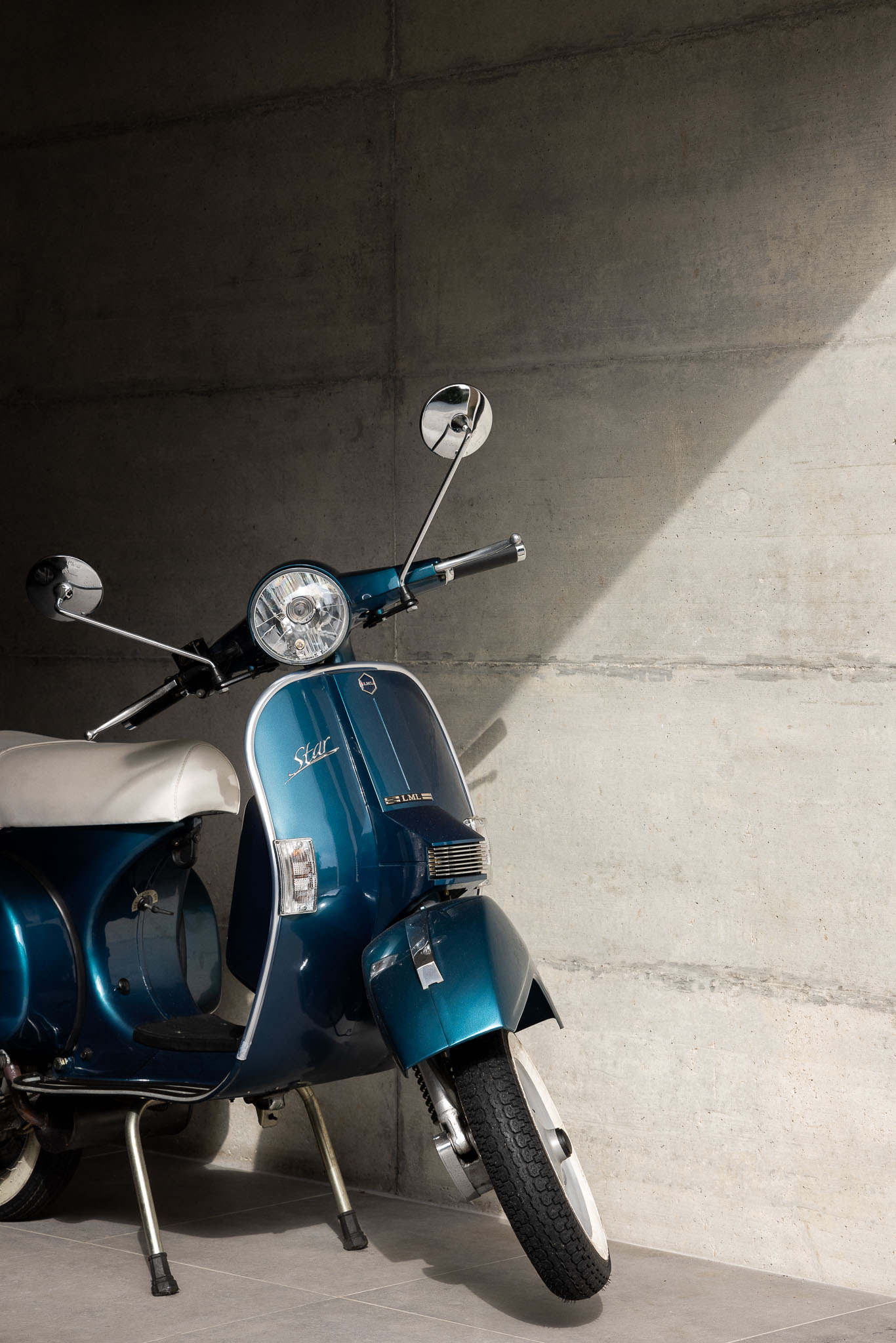 Vespa azul clássica estacionada em garagem com parede de betão aparente e piso cerâmico cinzento. — Casa H1 arquitetura Bruno H Gomes Arquitectura em São João da Madeira