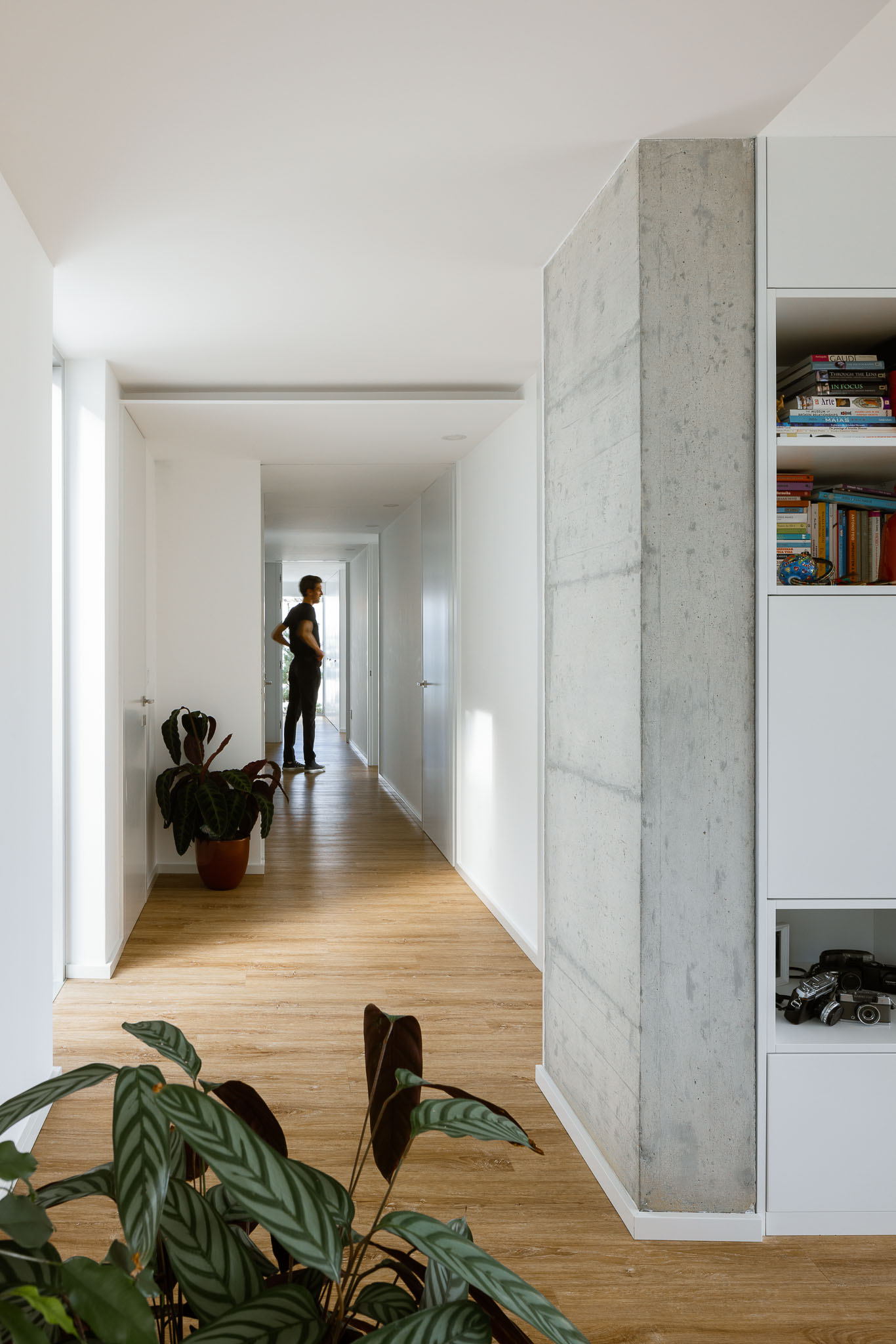 Corredor comprido com pavimento em madeira, paredes brancas, pilar em betão e estante com livros. — Casa H1 arquitetura Bruno H Gomes Arquitectura em São João da Madeira