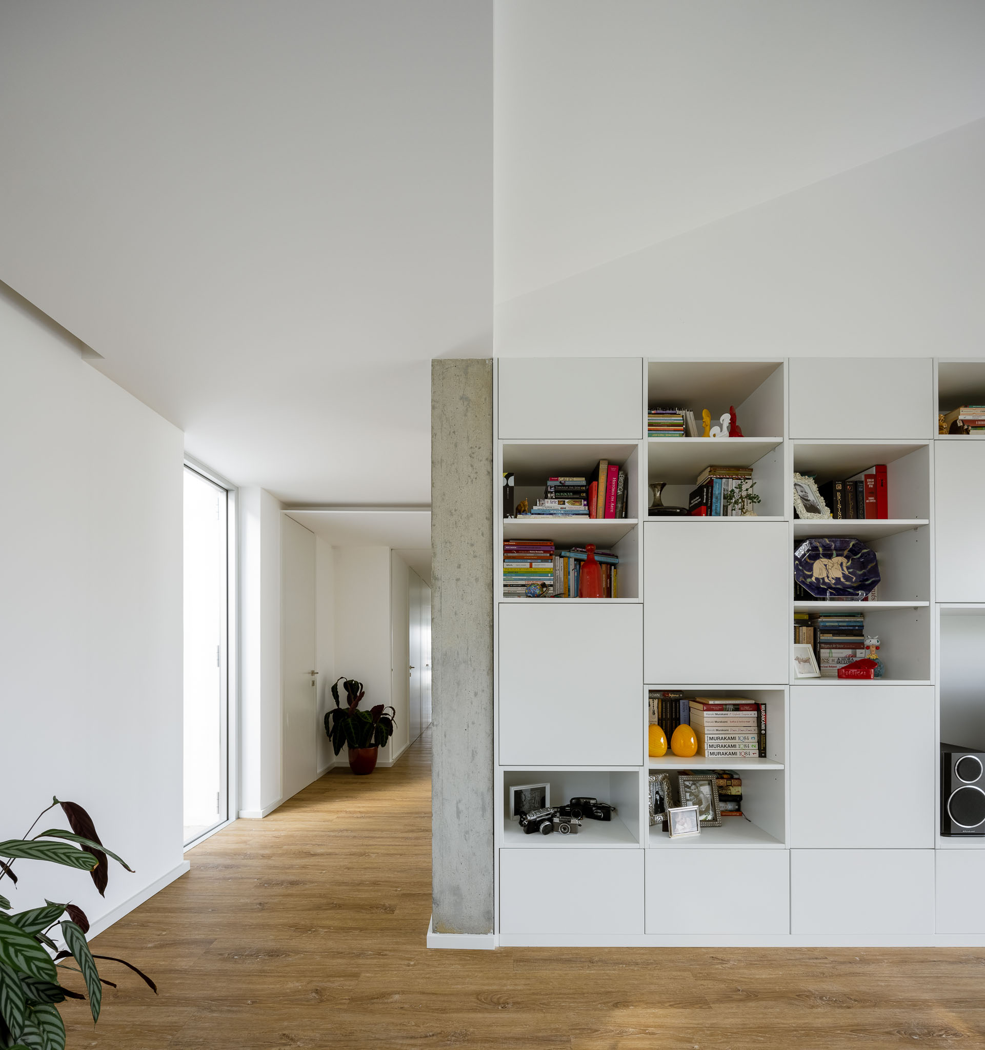 Sala de estar com estante branca modular, pilar em betão aparente e corredor em madeira ao fundo. — Casa H1 arquitetura Bruno H Gomes Arquitectura em São João da Madeira