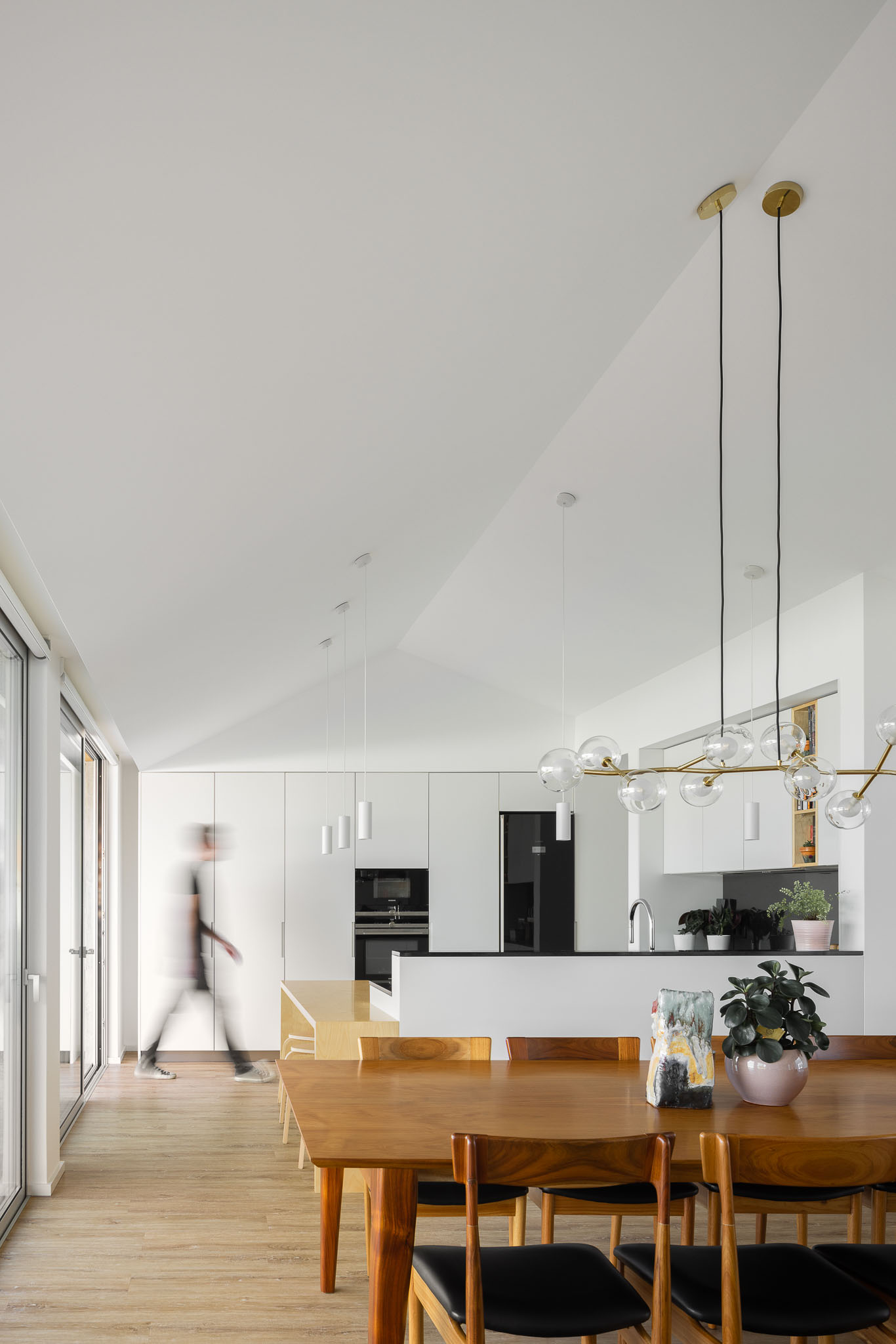 Sala de jantar e cozinha com mesa de madeira, cadeiras pretas, candeeiros suspensos e armários brancos. — Casa H1 arquitetura Bruno H Gomes Arquitectura em São João da Madeira