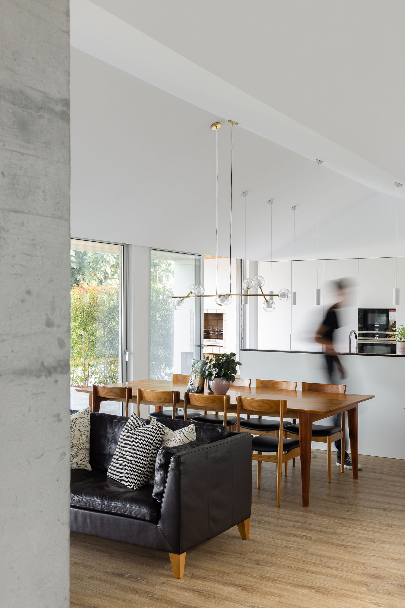Sala de estar e jantar com sofá de couro preto, mesa de madeira, cozinha branca e pilar de betão. — Casa H1 arquitetura Bruno H Gomes Arquitectura em São João da Madeira
