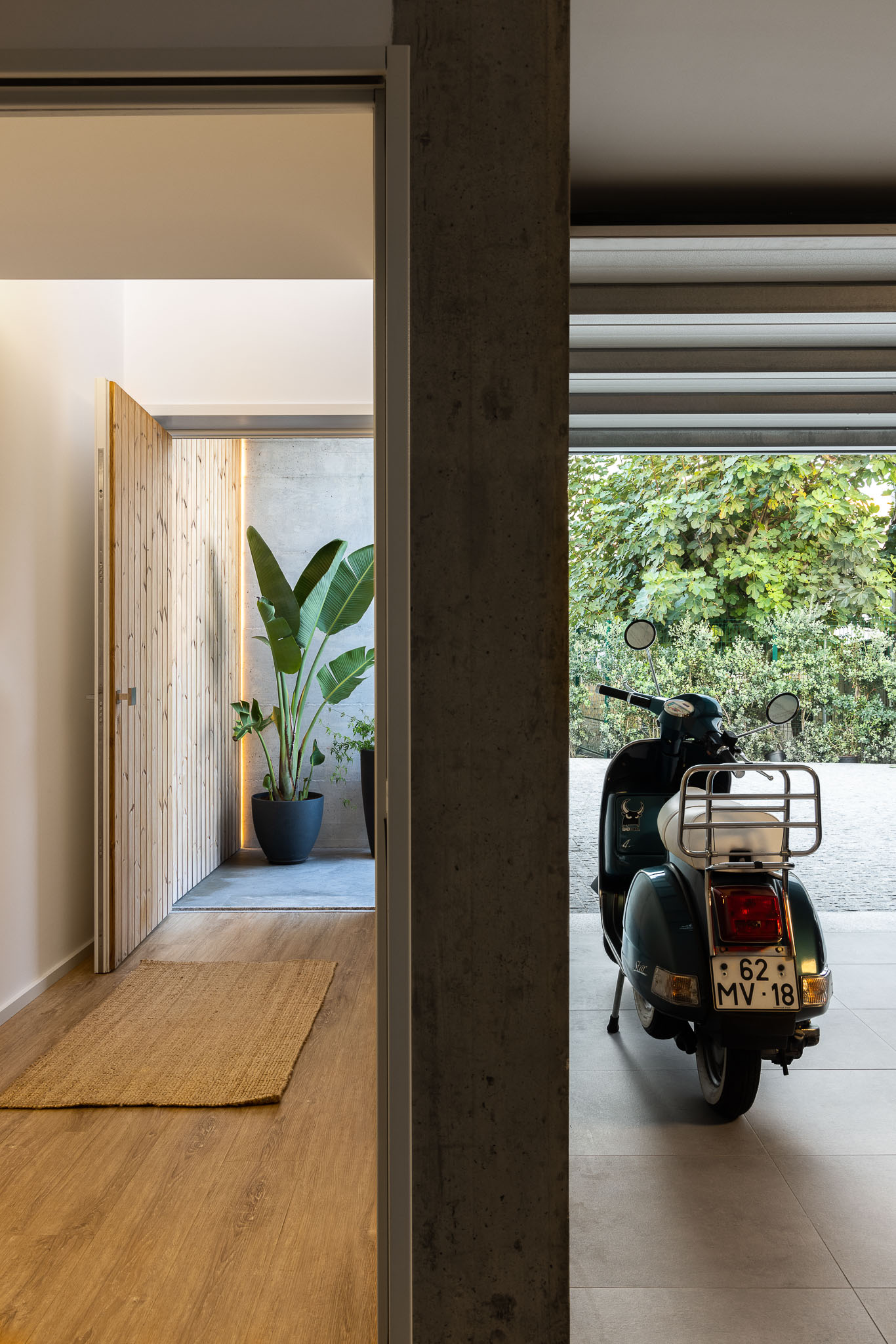 Hall de entrada com porta de madeira, tapete de juta, pilar de betão e garagem com Vespa ao fundo. — Casa H1 arquitetura Bruno H Gomes Arquitectura em São João da Madeira