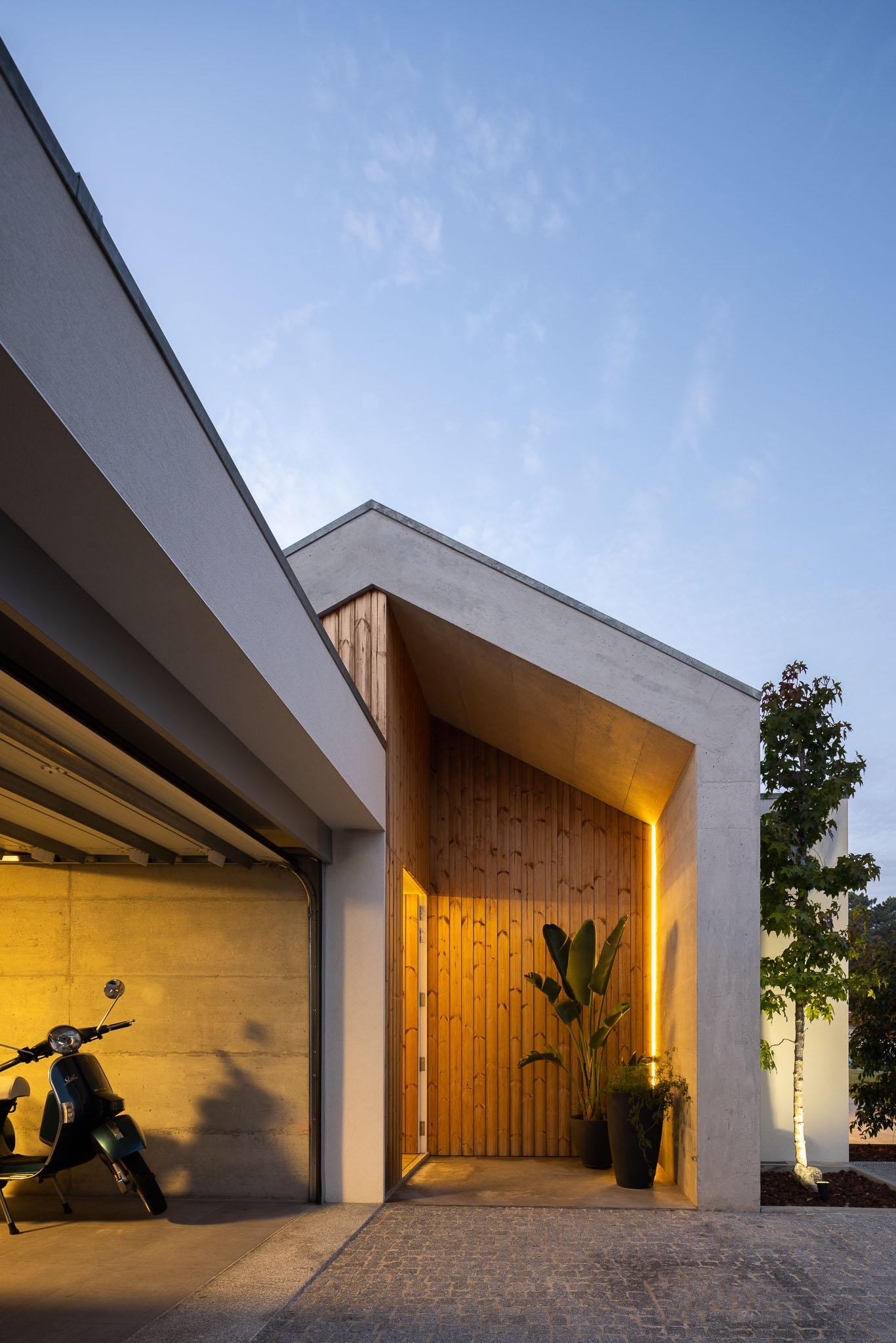 Entrada da casa iluminada ao anoitecer, com porta de madeira, garagem e Vespa. — Casa H1 arquitetura Bruno H Gomes Arquitectura em São João da Madeira