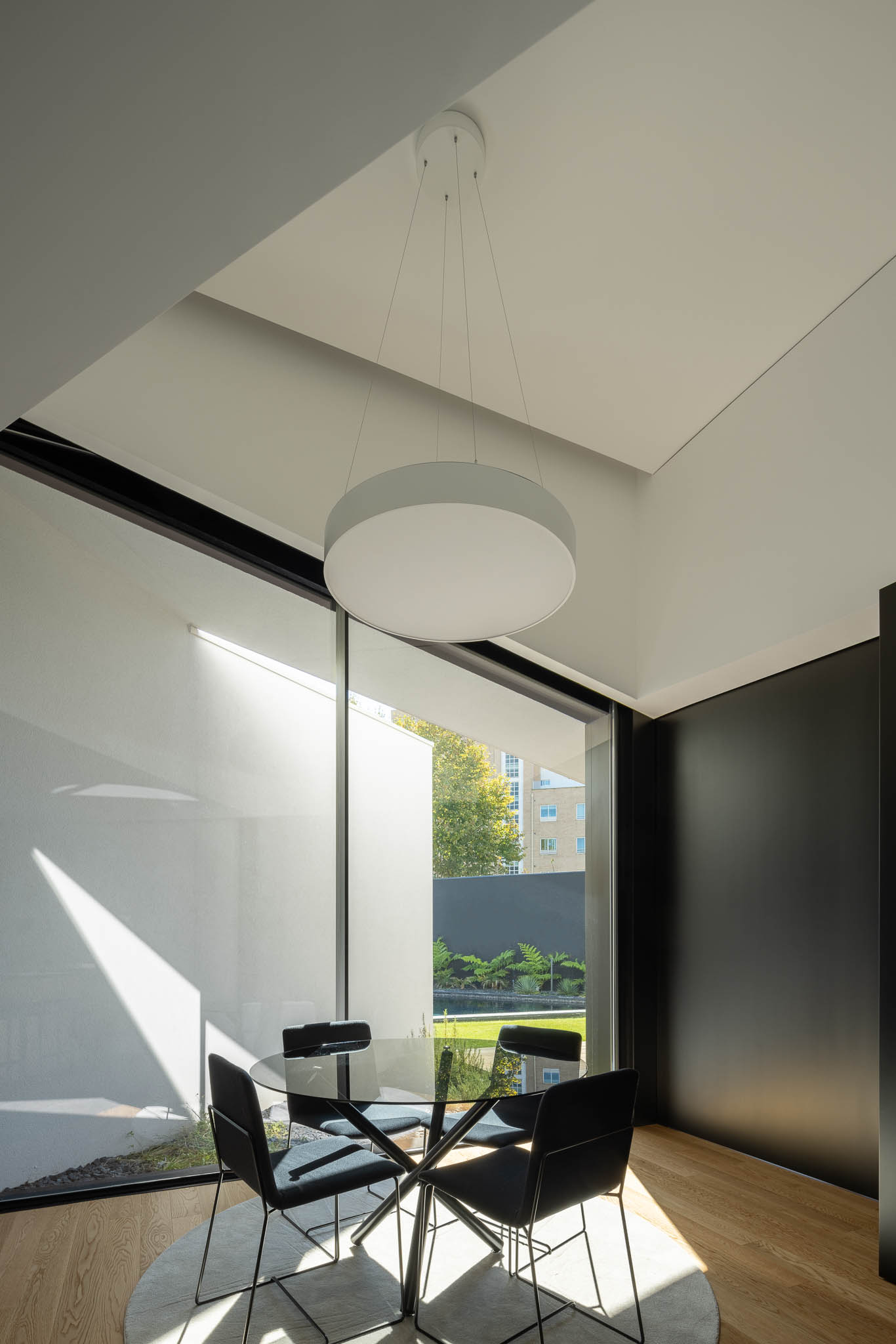 Sala de jantar com mesa redonda de vidro, cadeiras pretas e candeeiro suspenso circular, com luz natural. — Casa Tilt arquitetura Mutant Architecture and Design em Gondomar