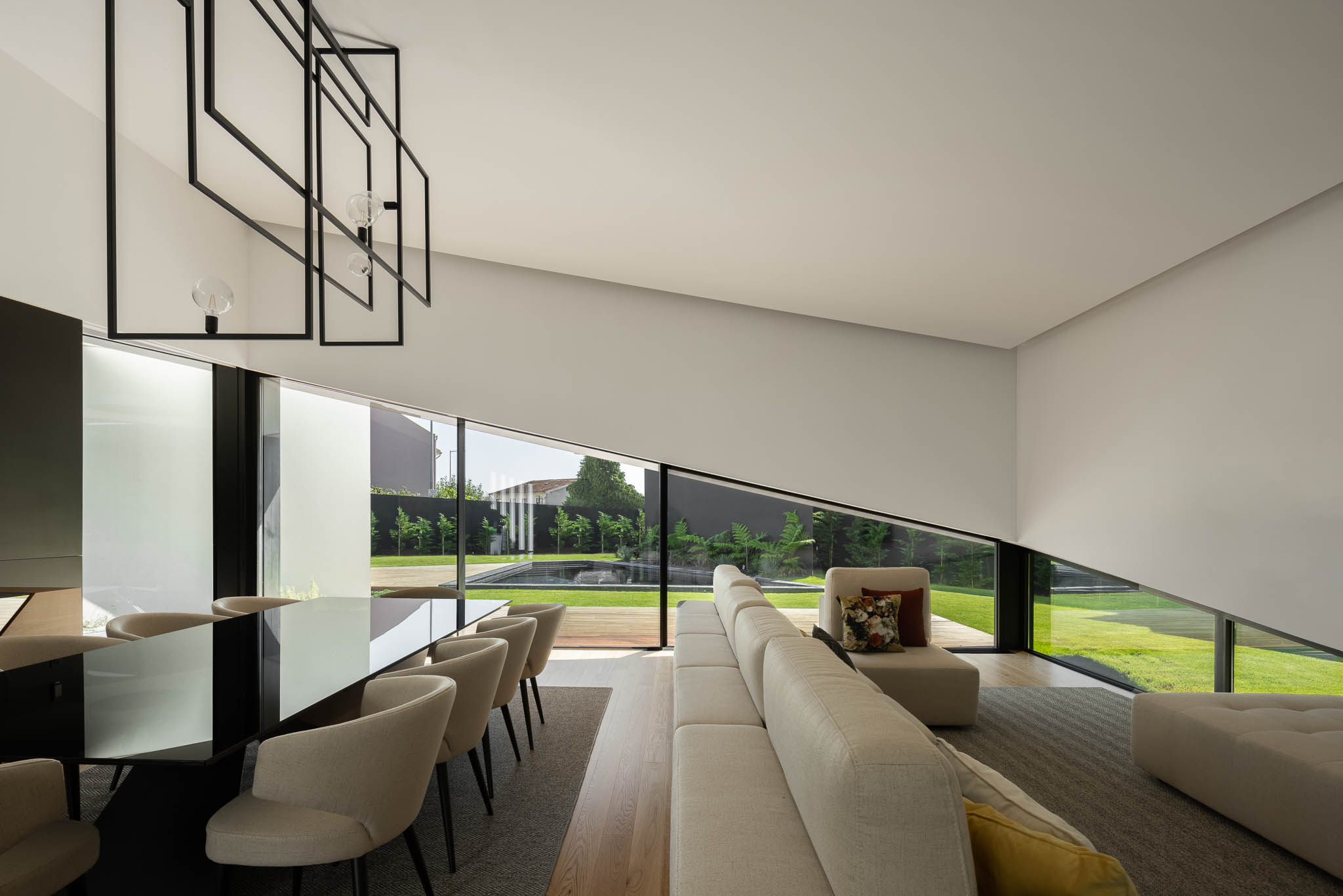Espaço amplo com sala de estar e jantar, janelas inclinadas, sofá bege e vista para o jardim com piscina. — Casa Tilt arquitetura Mutant Architecture and Design em Gondomar