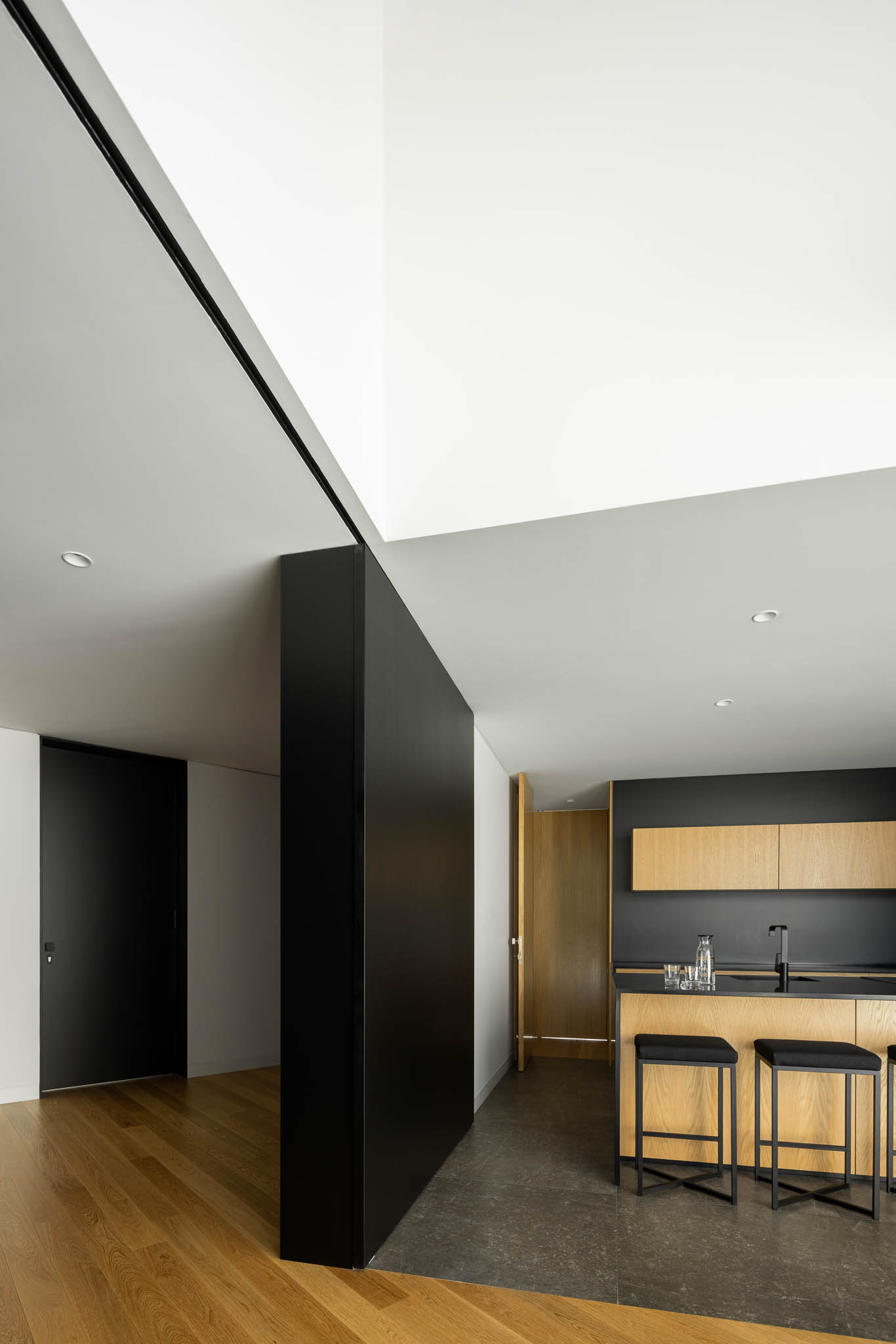 Divisória preta entre áreas, pavimento misto madeira/pedra, cozinha com armários de madeira e bancada preta. — Casa Tilt arquitetura Mutant Architecture and Design em Gondomar