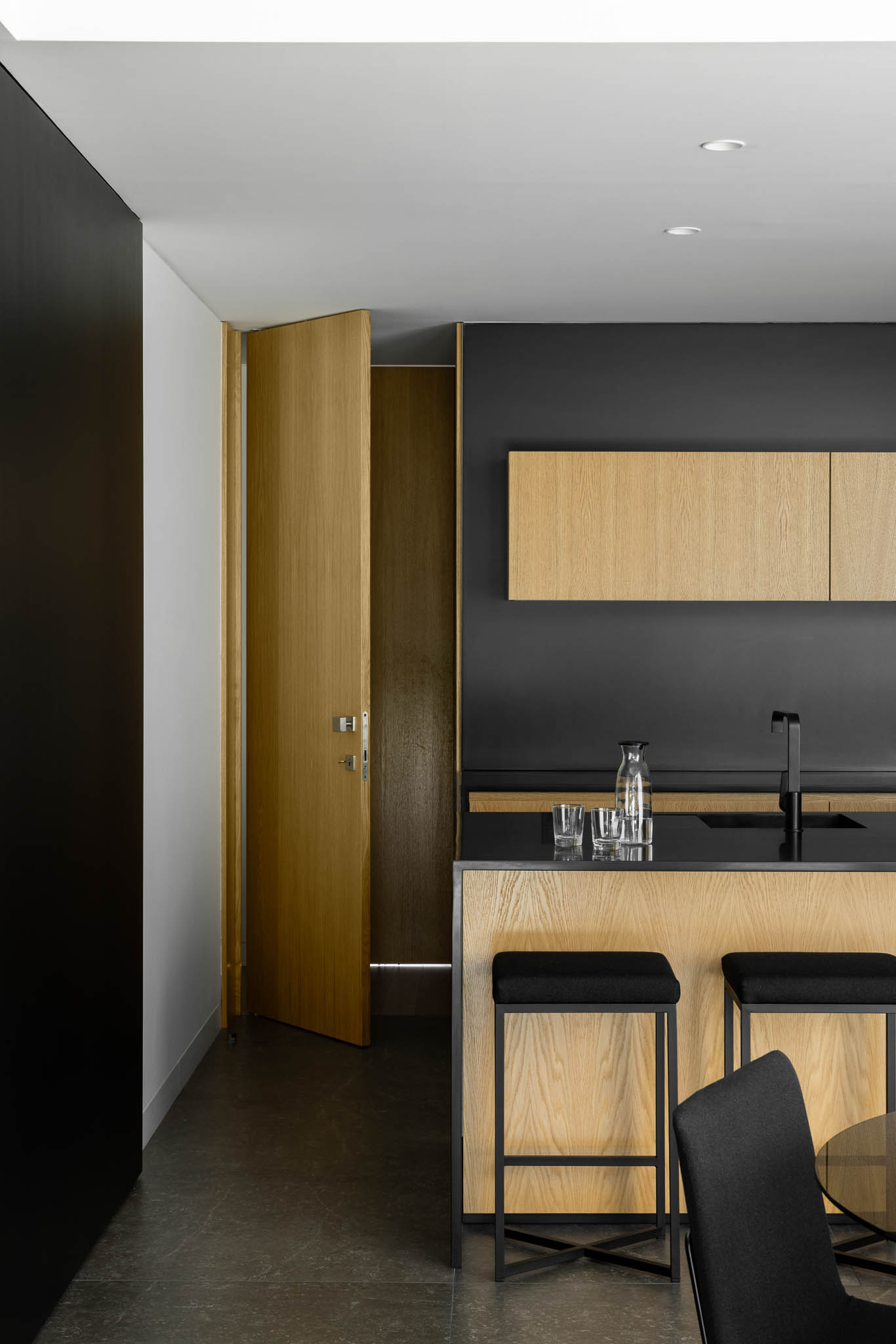 Detalhe da cozinha com armários em madeira clara, bancada preta, banquetas e porta aberta em madeira. — Casa Tilt arquitetura Mutant Architecture and Design em Gondomar