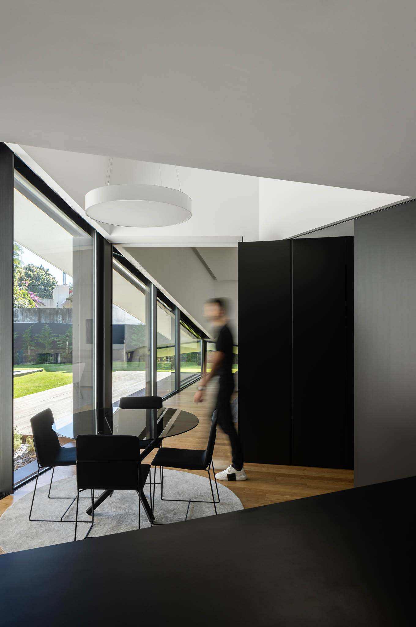 Sala de jantar com mesa redonda, cadeiras pretas, candeeiro suspenso e janelas amplas com vista para o jardim. — Casa Tilt arquitetura Mutant Architecture and Design em Gondomar