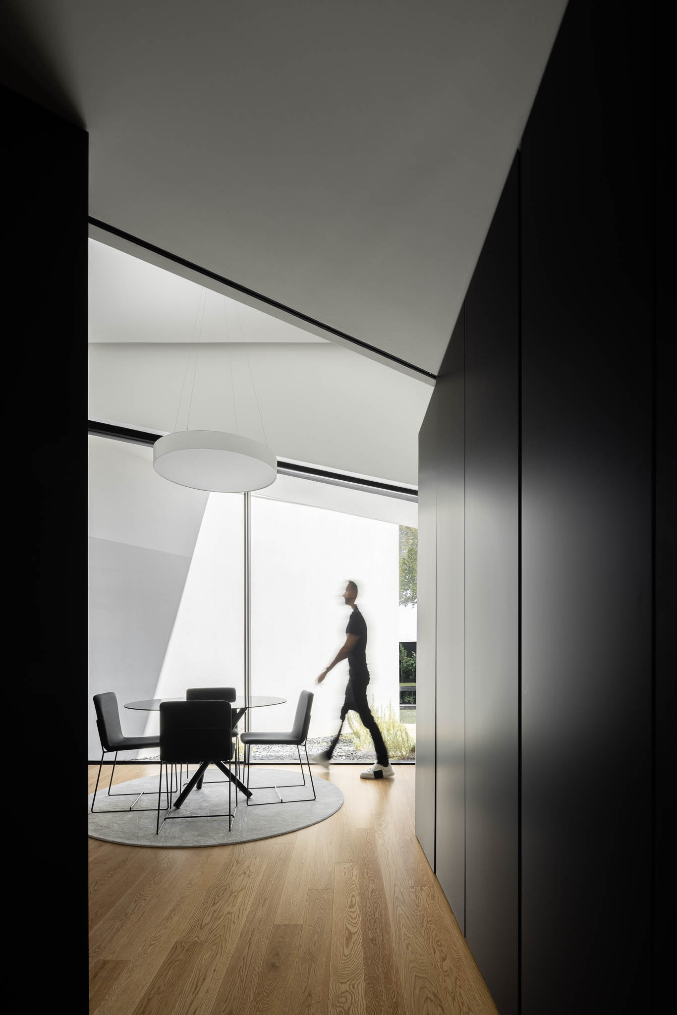 Homem a caminhar numa sala de jantar minimalista com mesa redonda e cadeiras pretas, sob candeeiro circular. — Casa Tilt arquitetura Mutant Architecture and Design em Gondomar