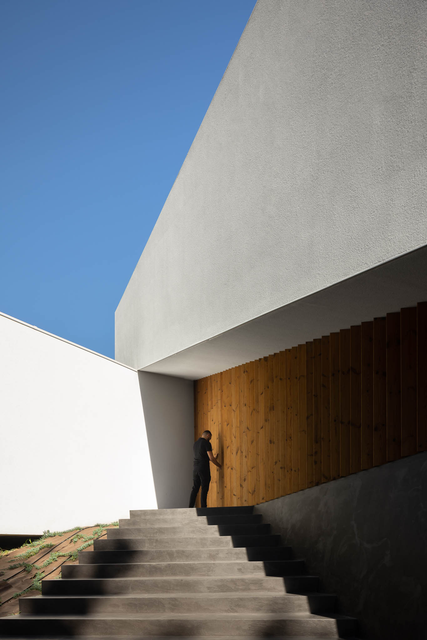 Escadaria exterior em betão cinzento, fachada branca com detalhe em ripas de madeira e homem a entrar. — Casa Tilt arquitetura Mutant Architecture and Design em Gondomar