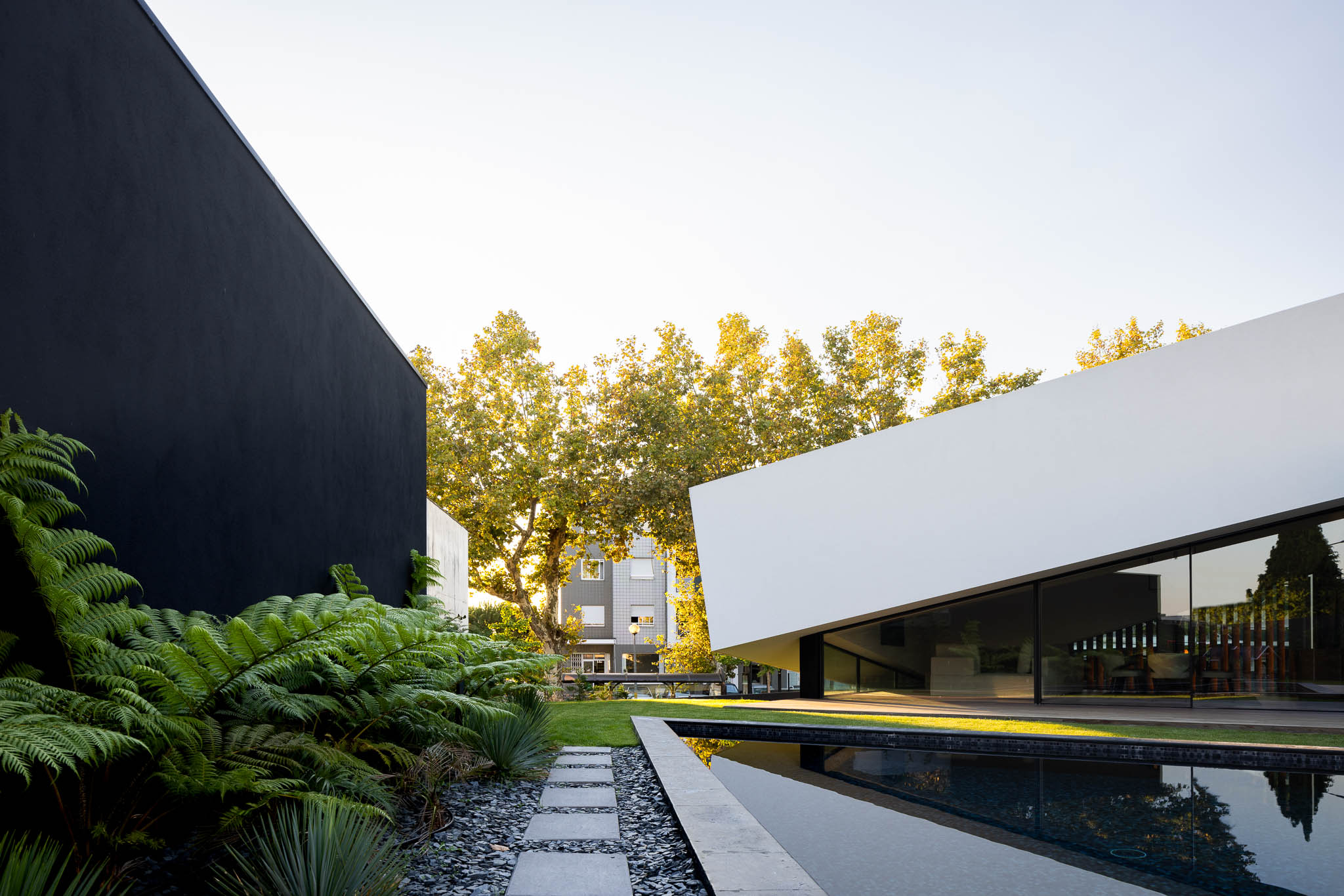Jardim com piscina, vegetação luxuriante e fachada branca angular com janelas, contrastando com parede preta. — Casa Tilt arquitetura Mutant Architecture and Design em Gondomar
