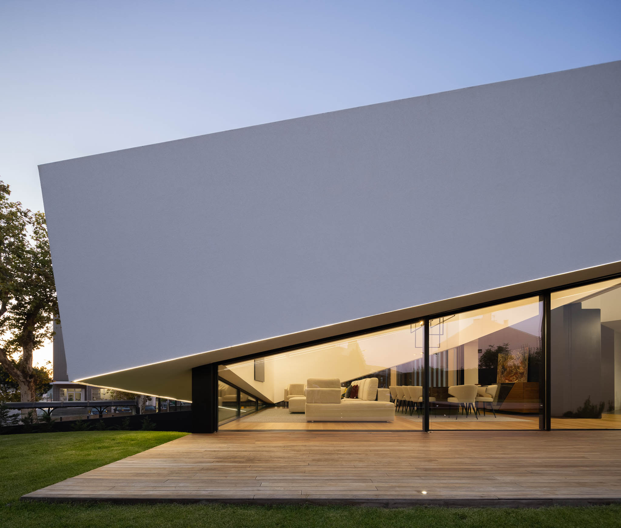 Fachada branca inclinada com janelas amplas, revelando interior iluminado e deck de madeira adjacente. — Casa Tilt arquitetura Mutant Architecture and Design em Gondomar