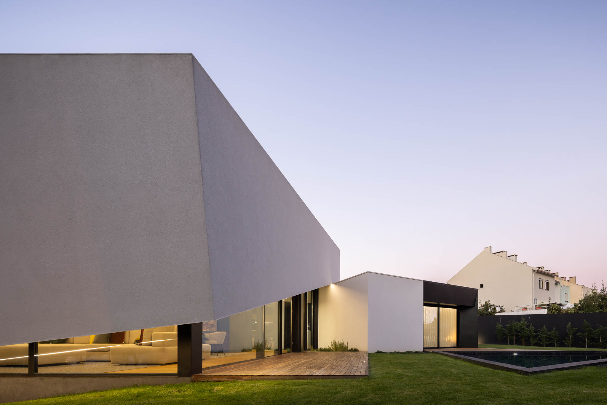 Vista exterior da casa com fachada branca angular, deck de madeira, piscina e relvado ao entardecer. — Casa Tilt arquitetura Mutant Architecture and Design em Gondomar