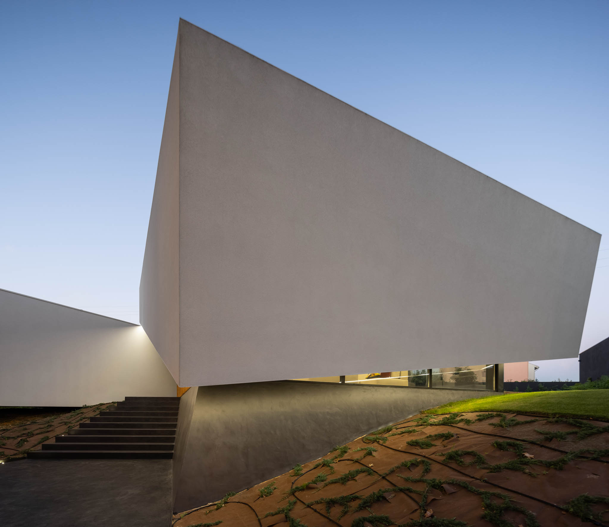 Fachada branca angular com escadaria em betão, vegetação rasteira e iluminação indireta sob a estrutura. — Casa Tilt arquitetura Mutant Architecture and Design em Gondomar