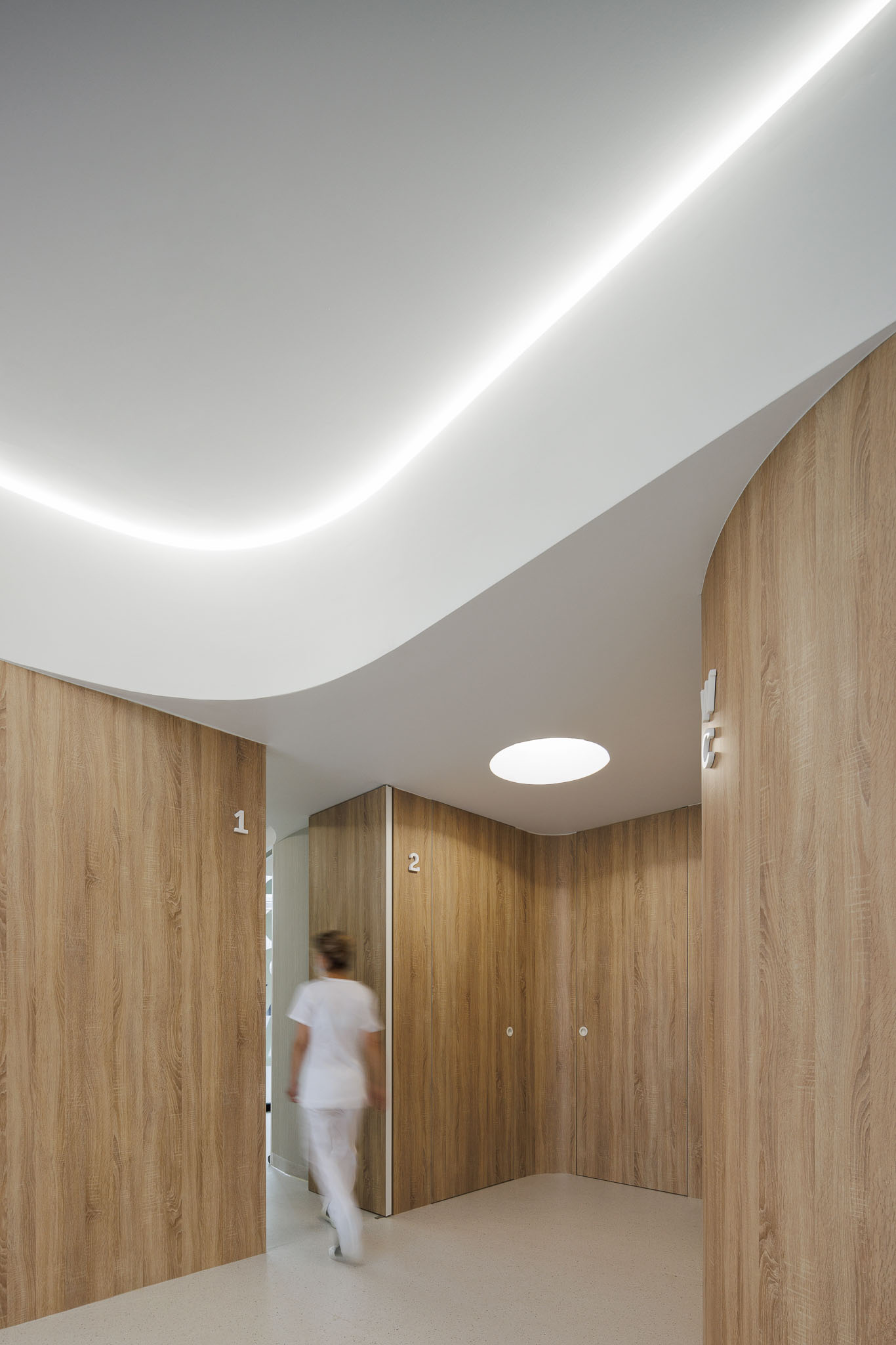 Corredor com portas numeradas em madeira clara, iluminação embutida e teto curvo branco. — MOOD Medicina Dentária arquitetura Tsou Arquitectos em Maia