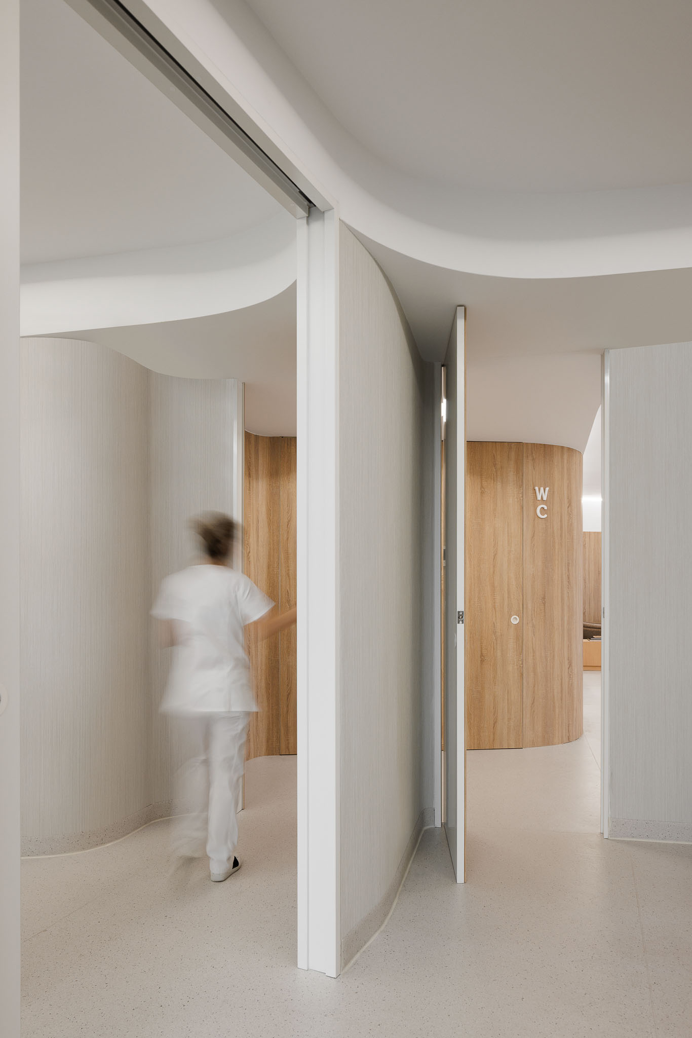 Corredor com paredes curvas revestidas a branco e madeira, porta aberta para WC. — MOOD Medicina Dentária arquitetura Tsou Arquitectos em Maia