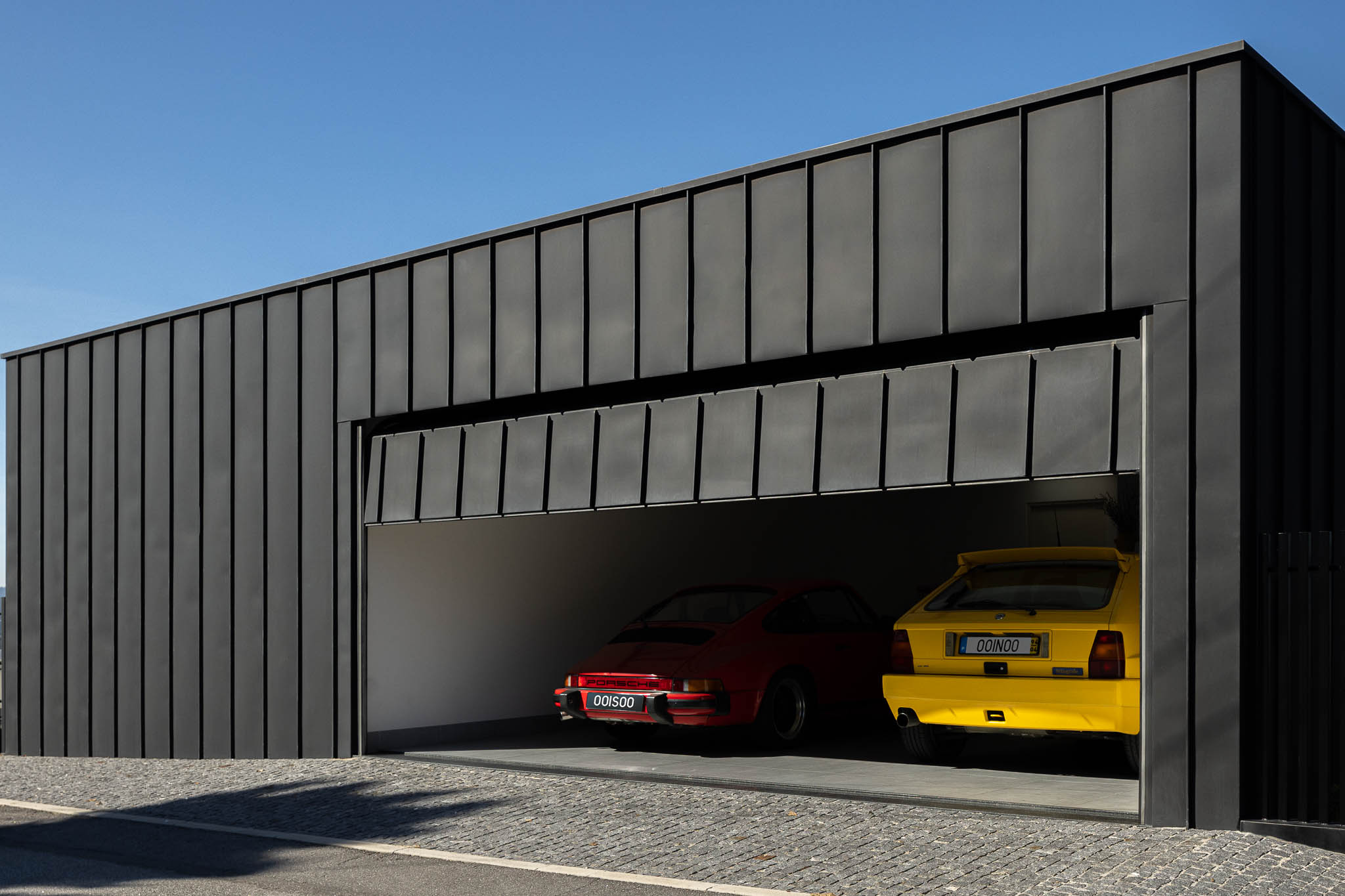 Garagem com revestimento em zinco preto, porta seccionada aberta e dois carros clássicos no interior. — Casa PS arquitetura Inception Architects Studio em Braga